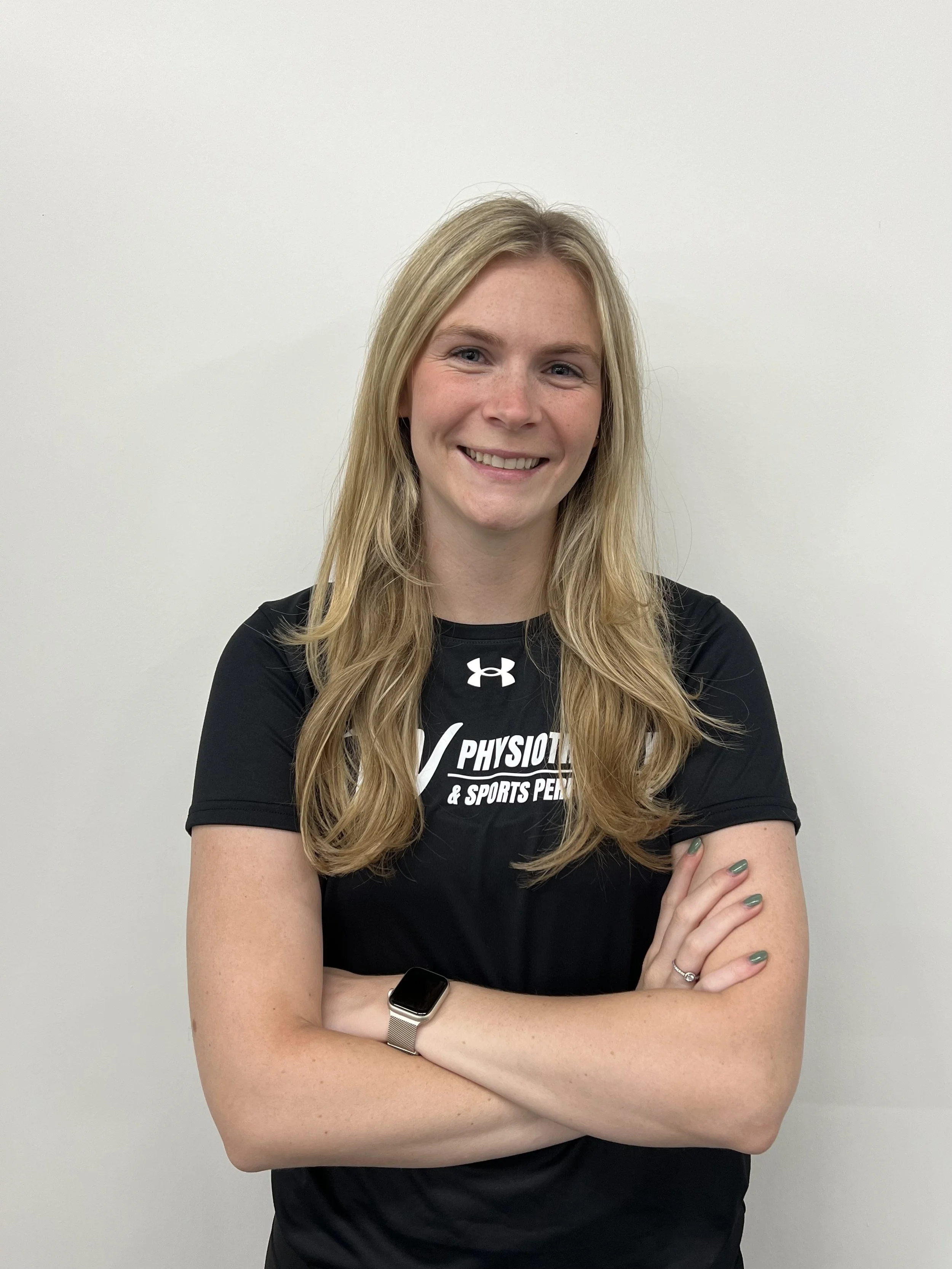 A woman with long blonde hair smiling, wearing a black Under Armour T-shirt with 'W Physiotherapy & Sports Performance' printed on it, standing against a plain white wall.