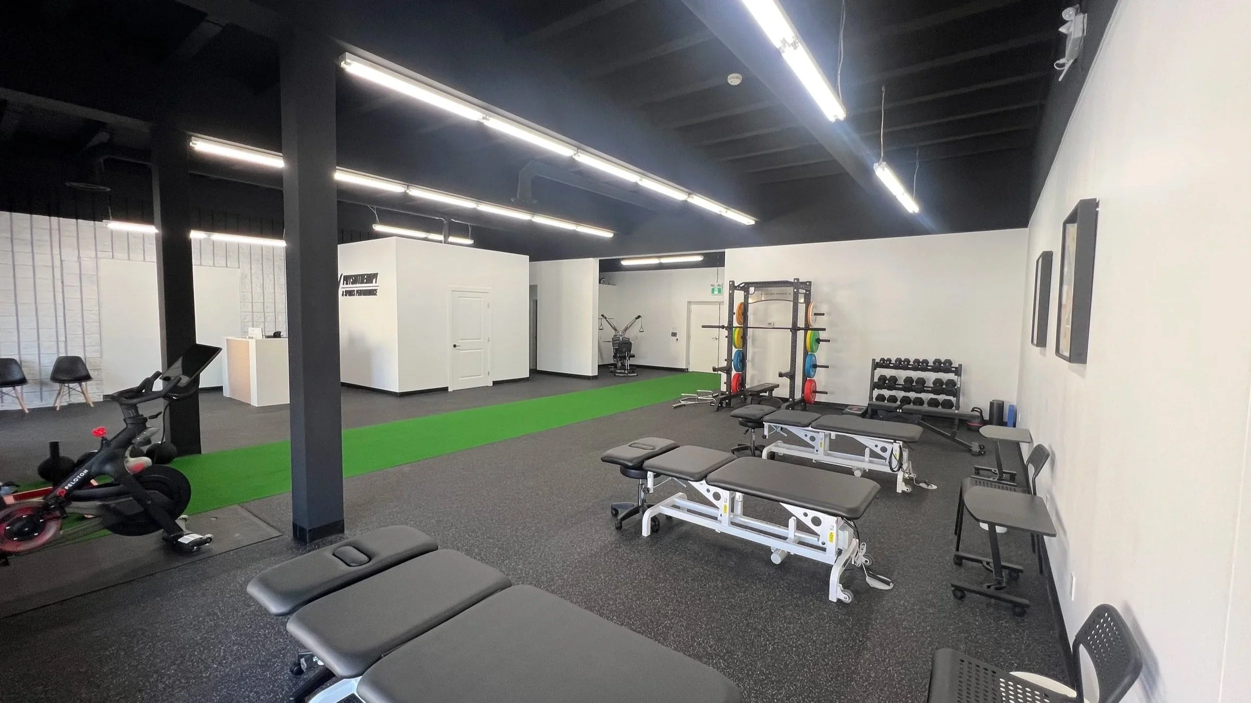 A gym with black ceiling and walls, brightly lit by long ceiling lights. There are workout benches, a stationary bike, kettlebells, dumbbells, and weight plates arranged on racks, along with an artificial turf strip on the black rubber mat floor.