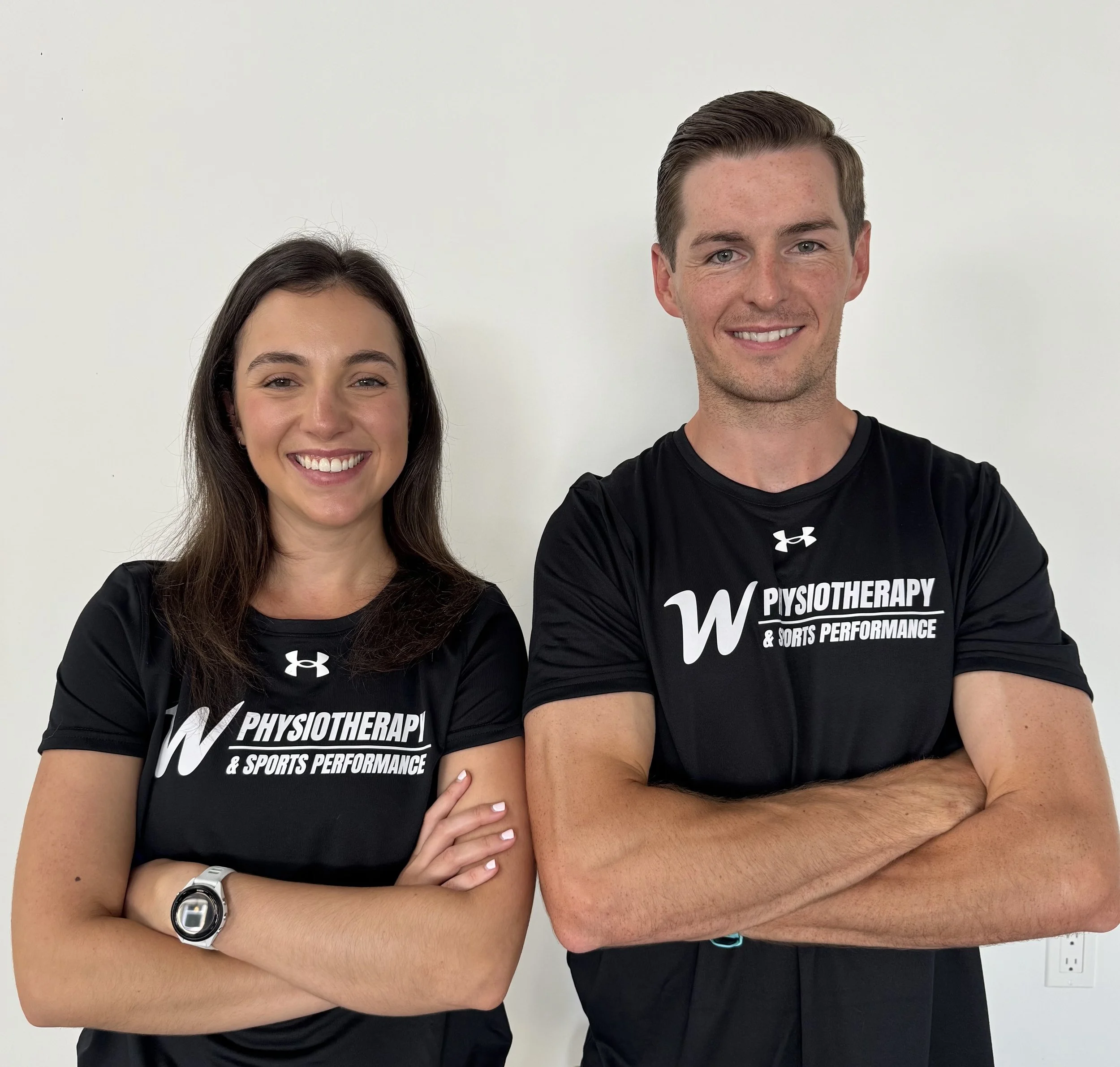 A woman and man wearing black T-shirts with the words 'Physiotherapy & Sports Performance' standing against a plain white wall, both smiling and crossing their arms.