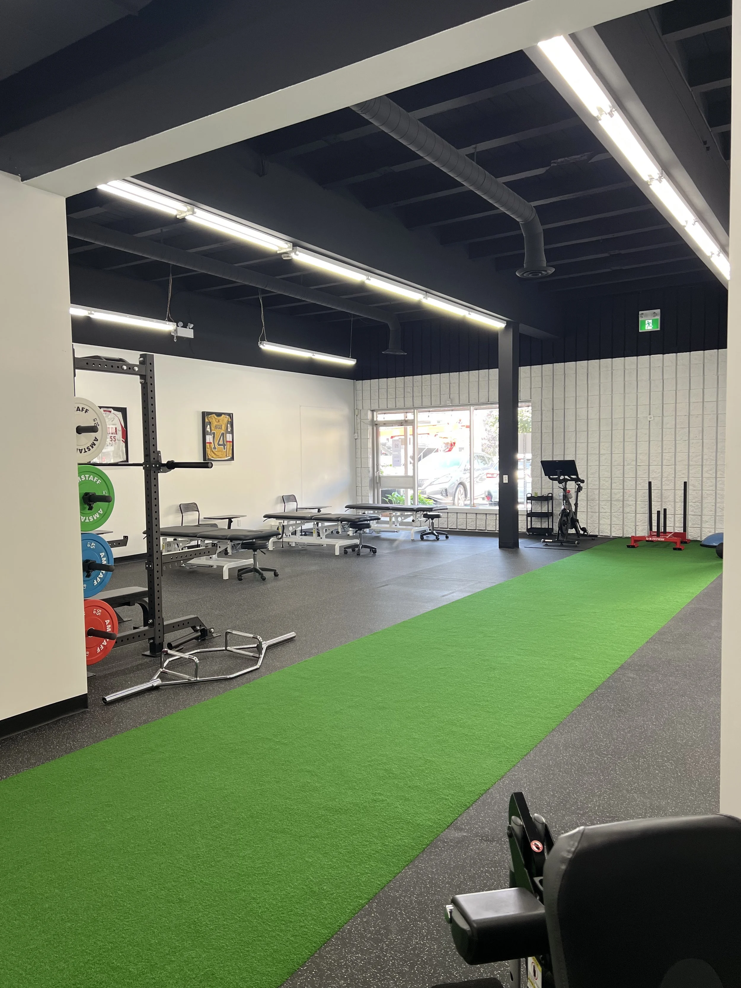 Interior of a gym with weightlifting equipment, benches, a treadmill, artificial turf, and a large window with cars outside.