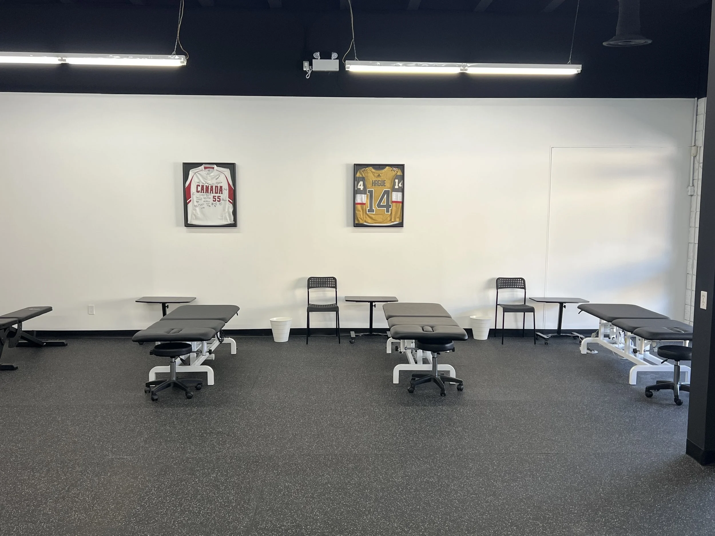 A room with three treatment tables lined up, each accompanied by a chair and a small table, with framed hockey jerseys on the white wall behind them.