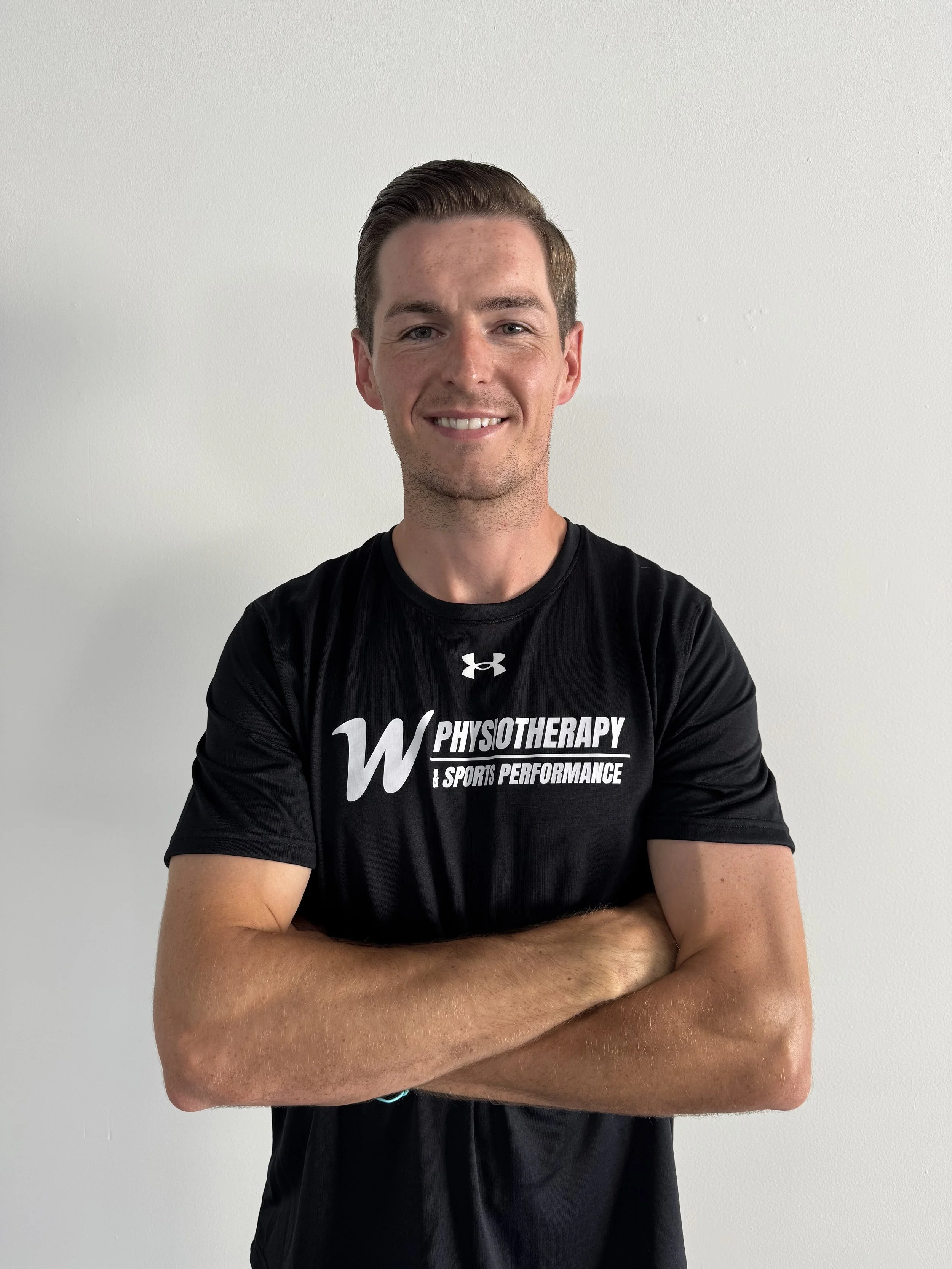 A young man with short brown hair and fair skin, smiling confidently with arms crossed, wearing a black athletic T-shirt that says 'Physiotherapy & Sports Performance' and a small Under Armour logo, standing against a plain white wall.