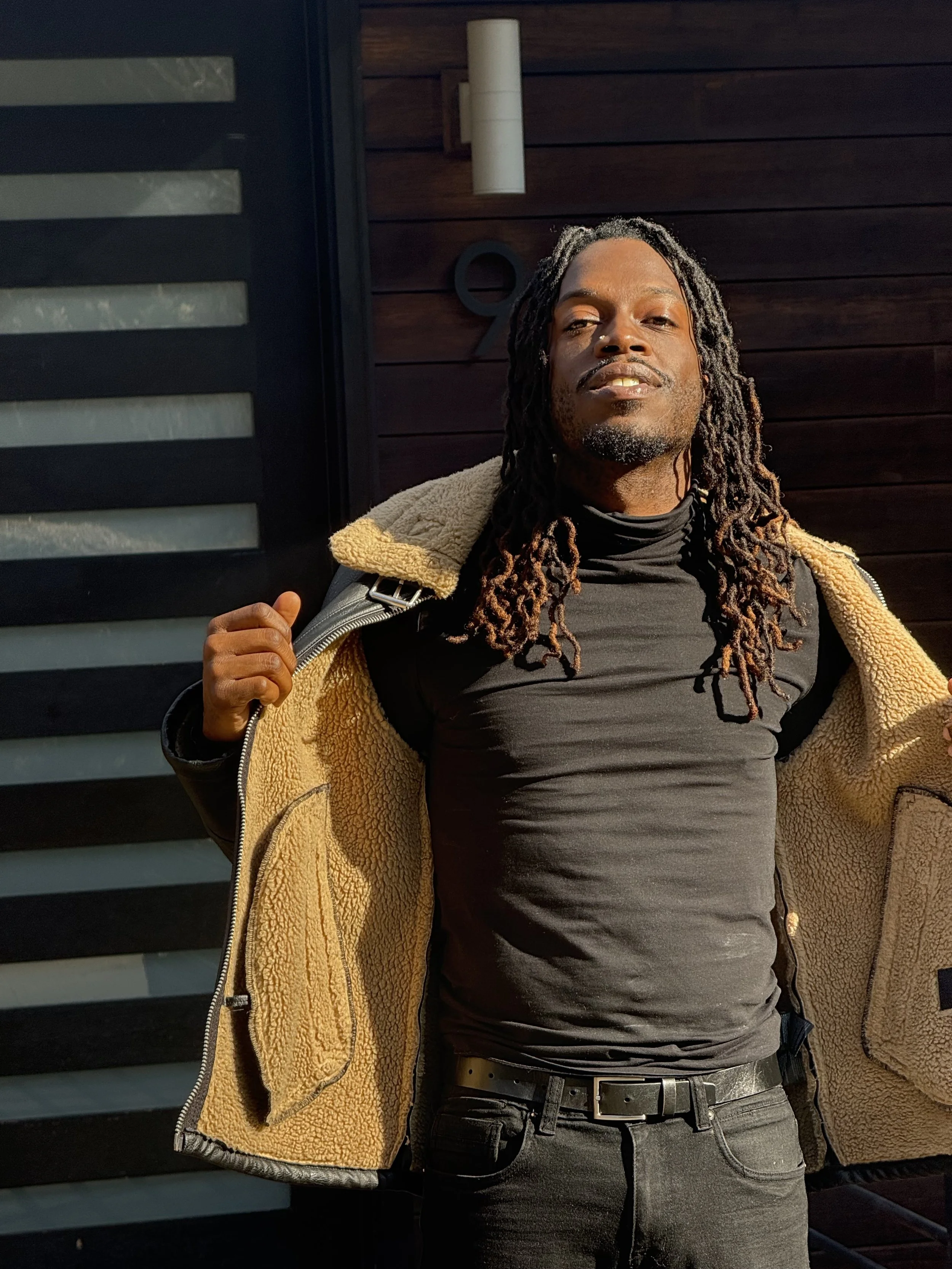 Young man with dreadlocks wearing a black shirt and black pants, opening a beige fleece jacket, standing outdoors in front of a dark wooden wall with a modern light fixture.