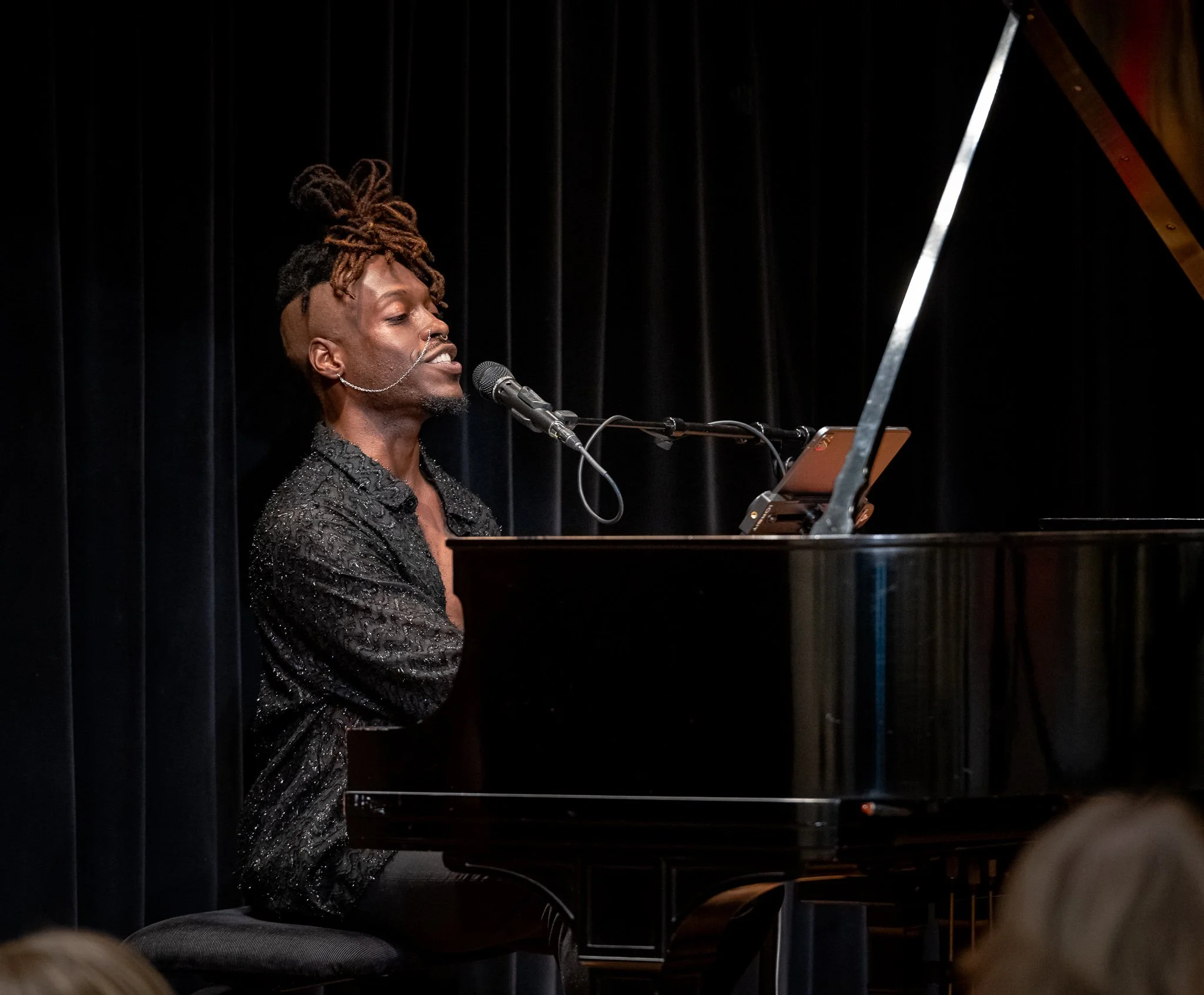 A man with dreadlocks styled in an elaborate updo, wearing a sparkly black shirt, is singing into a microphone while playing a grand piano on a stage with black curtains in the background.