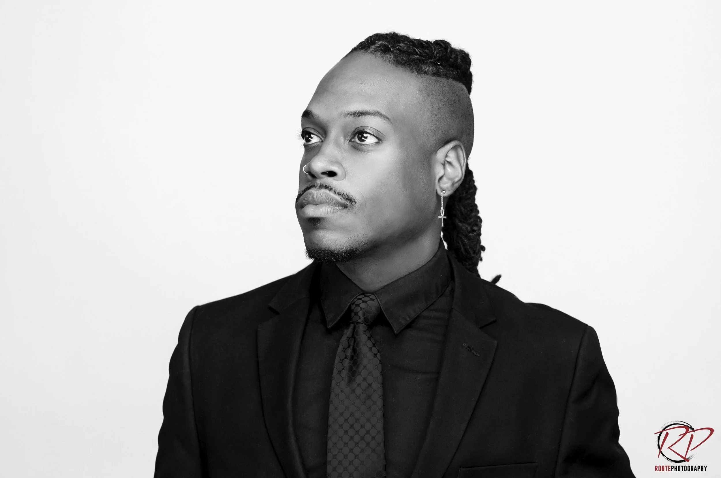 Black and white portrait of a man wearing a suit with a shirt and tie, with an earring, looking to the left, against a plain white background.