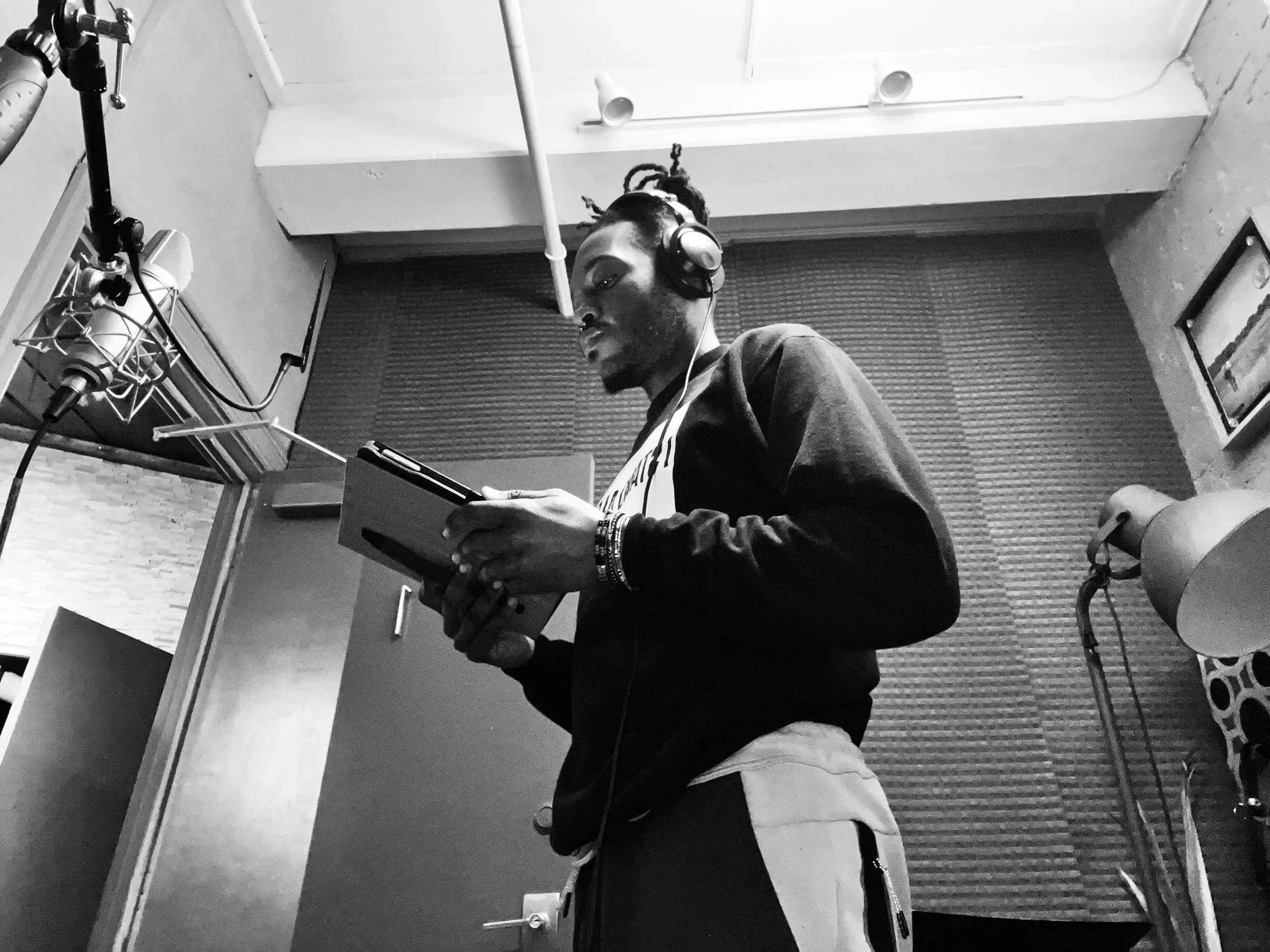 A man wearing headphones is standing in a recording studio, holding a tablet and looking at it.
