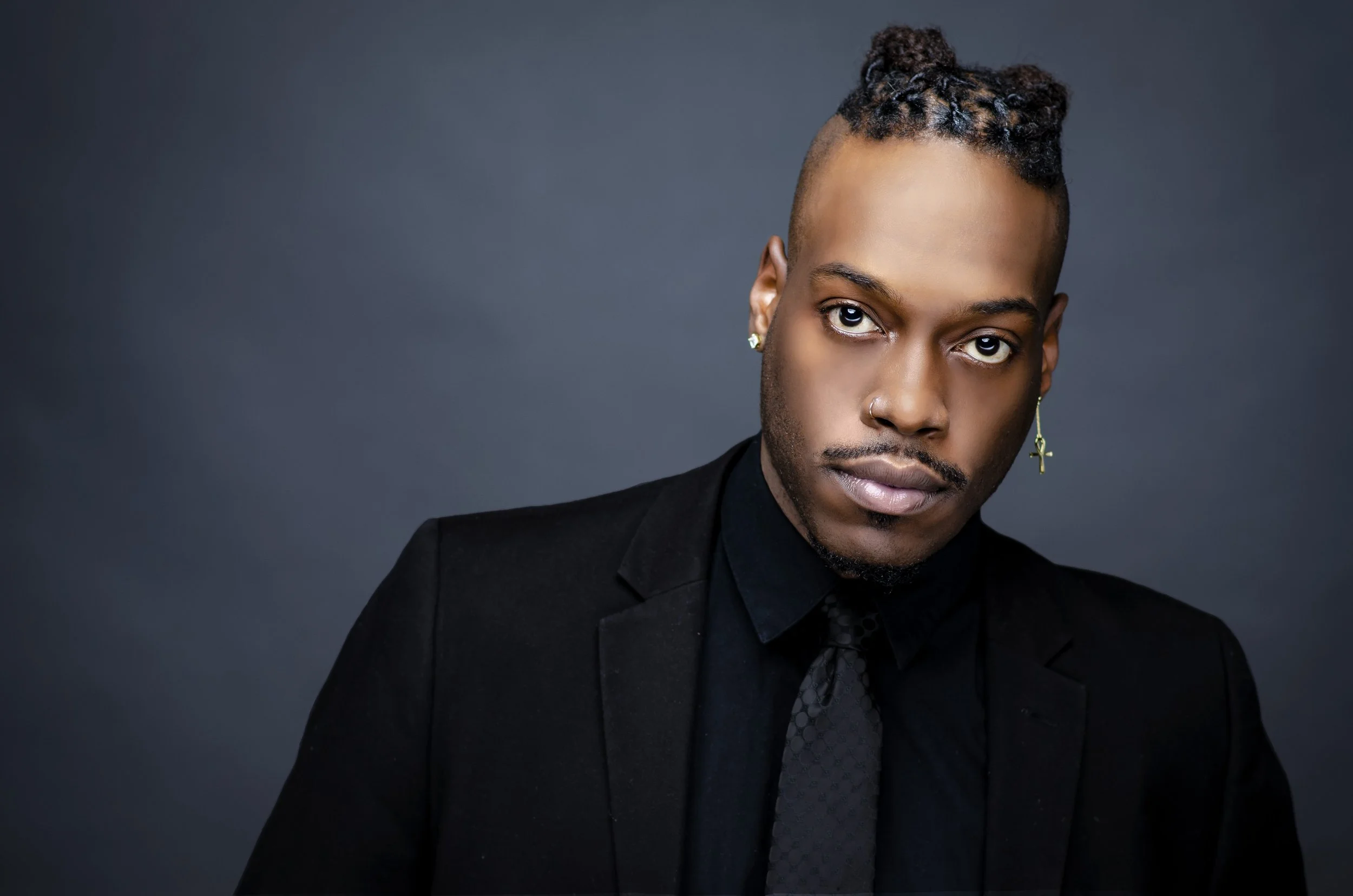 A young Black man with styled hair, wearing a black suit and tie, with earrings and a nose ring, posing against a dark gray background.