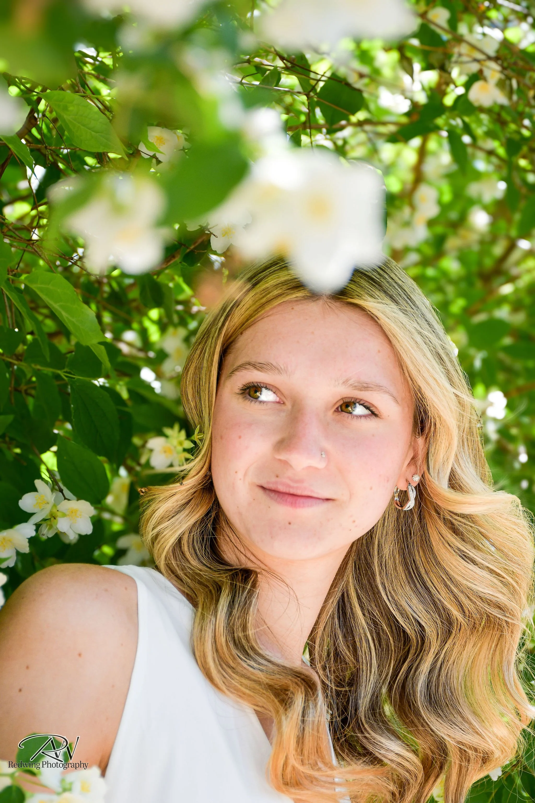 A young woman with blonde hair and earrings standing outdoors among green leaves and white flowers, looking to her right with a gentle smile.