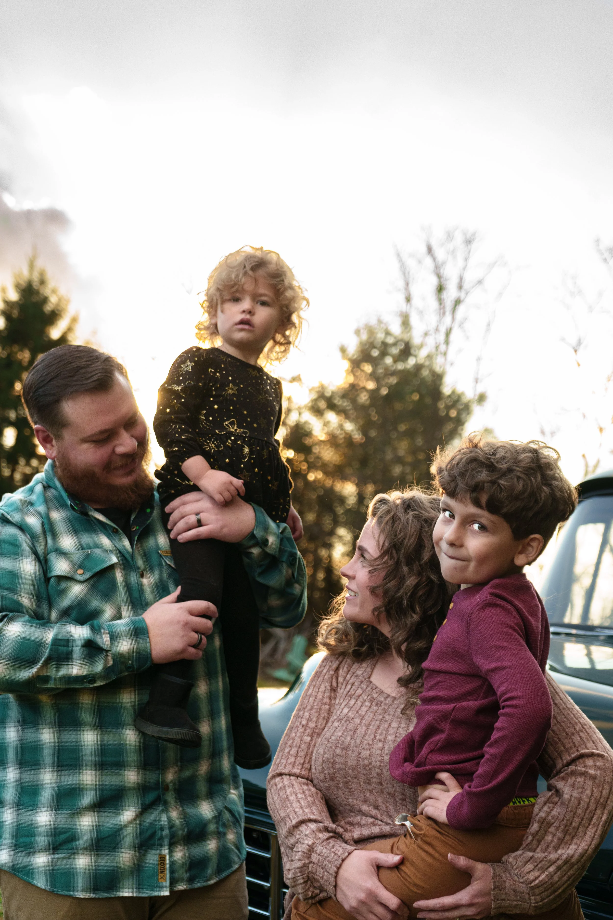 A family of four, including a man, woman, and two children, are outdoors near a black vehicle during sunset, with trees in the background. The man is holding a young girl on his shoulder, both are smiling. The woman is holding a boy, and they all app