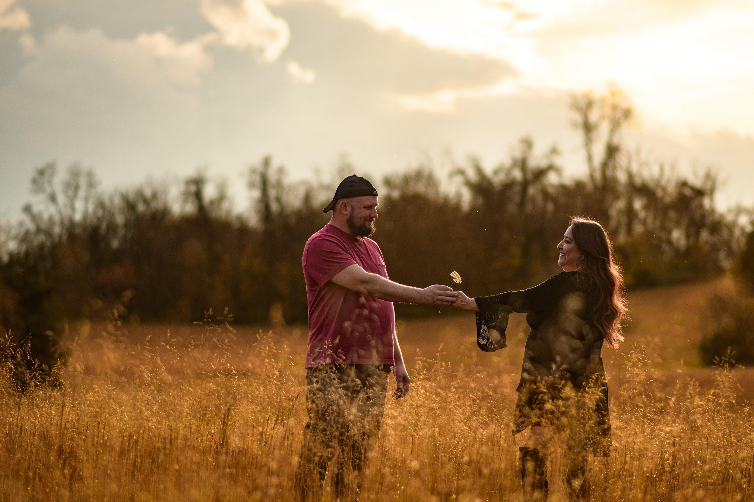 A man and woman standing in a field during sunset. The man is giving the woman a flower, and they are smiling at each other.