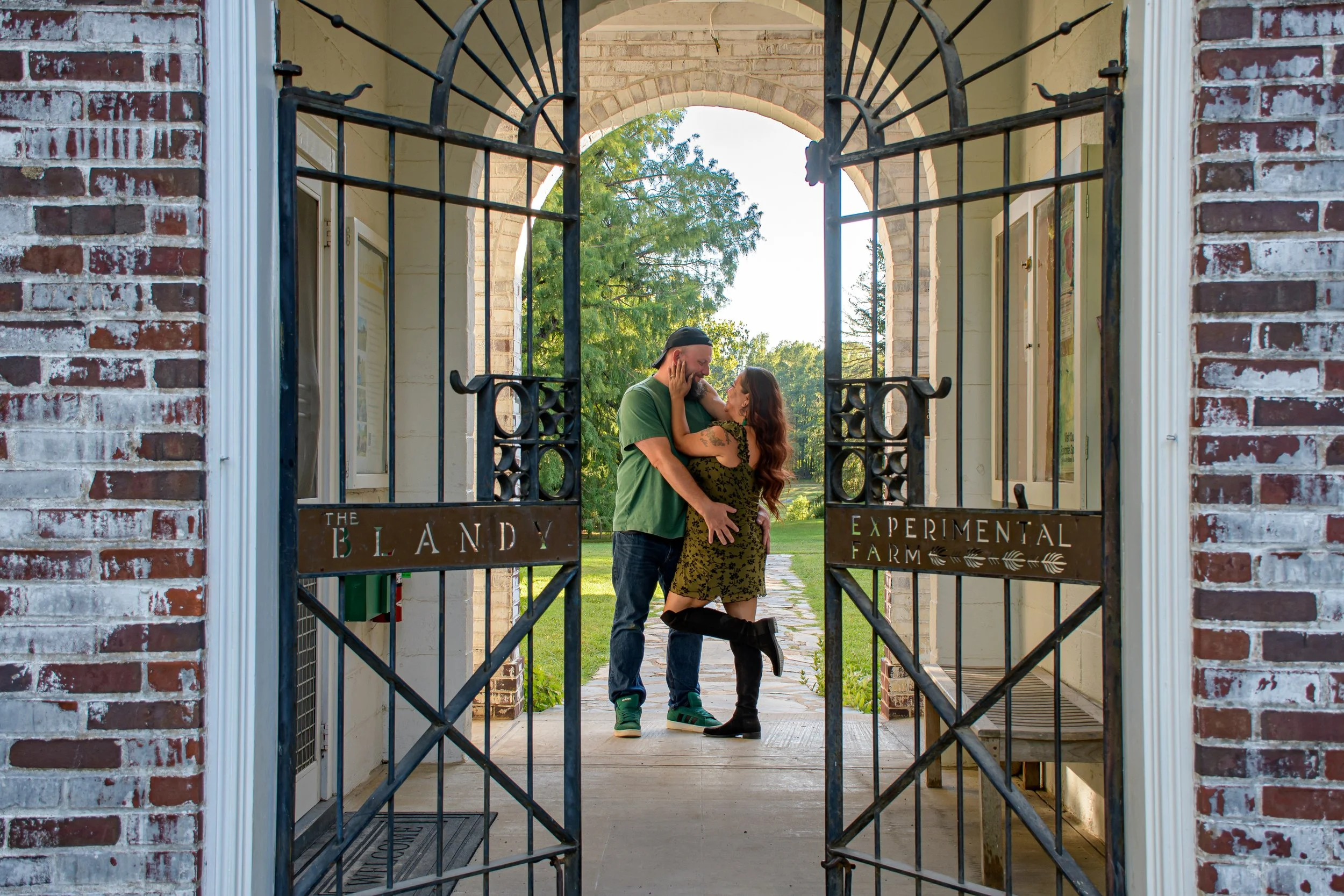 A man and woman sharing a kiss and laughter under a wrought iron gate with signs reading 'The Blandy' and 'Experimental Farm' in a park setting.