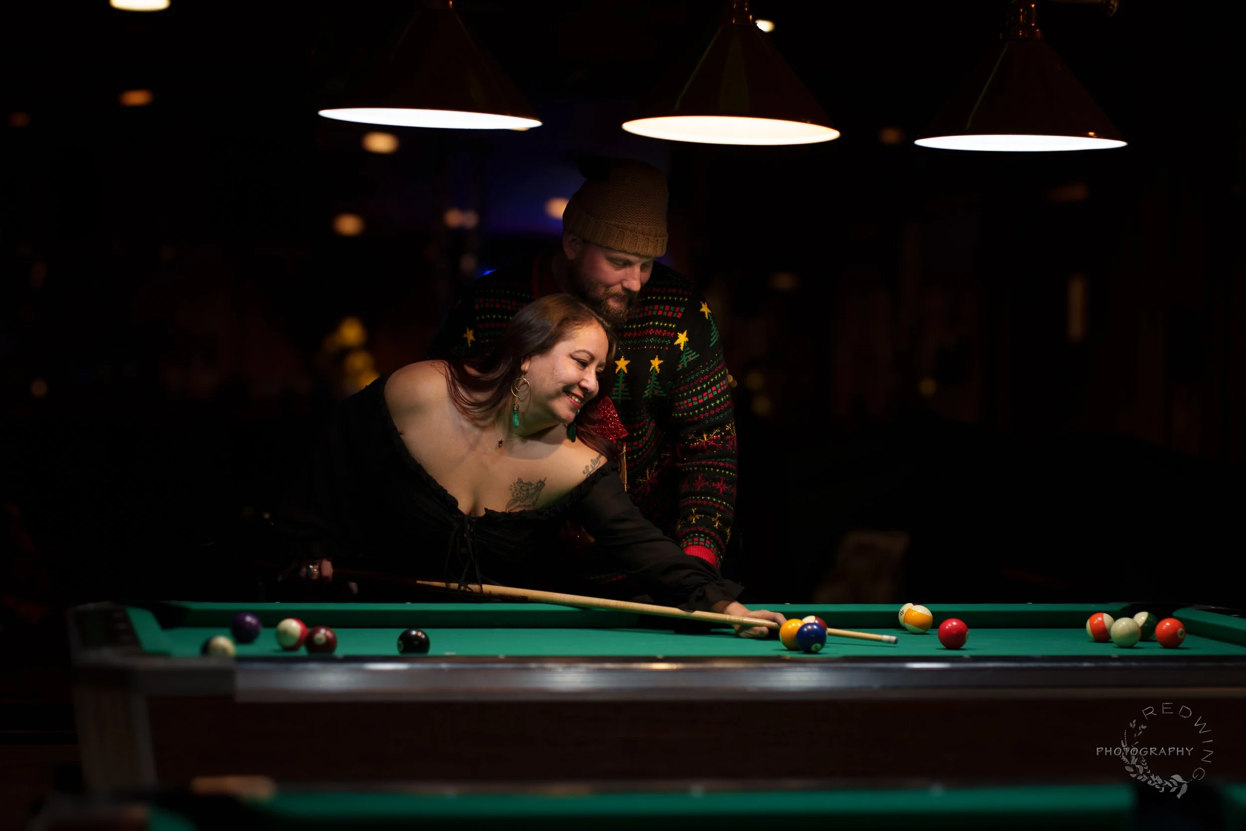 A man and woman playing pool together in a dimly lit bar, with the woman aiming to take a shot and both enjoying the moment.