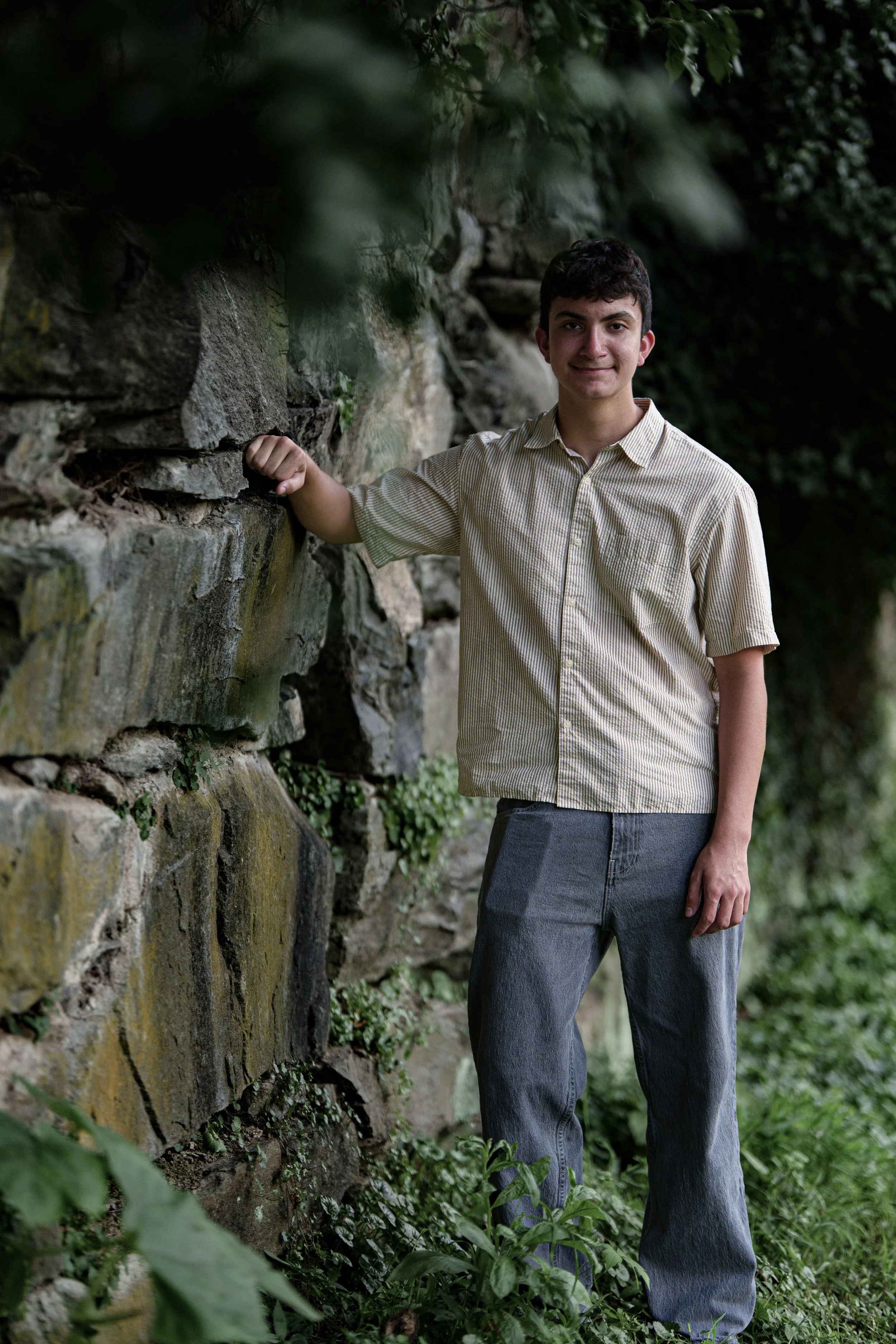 A young man wearing a beige striped shirt and gray jeans standing outdoors next to a stone wall with greenery around.