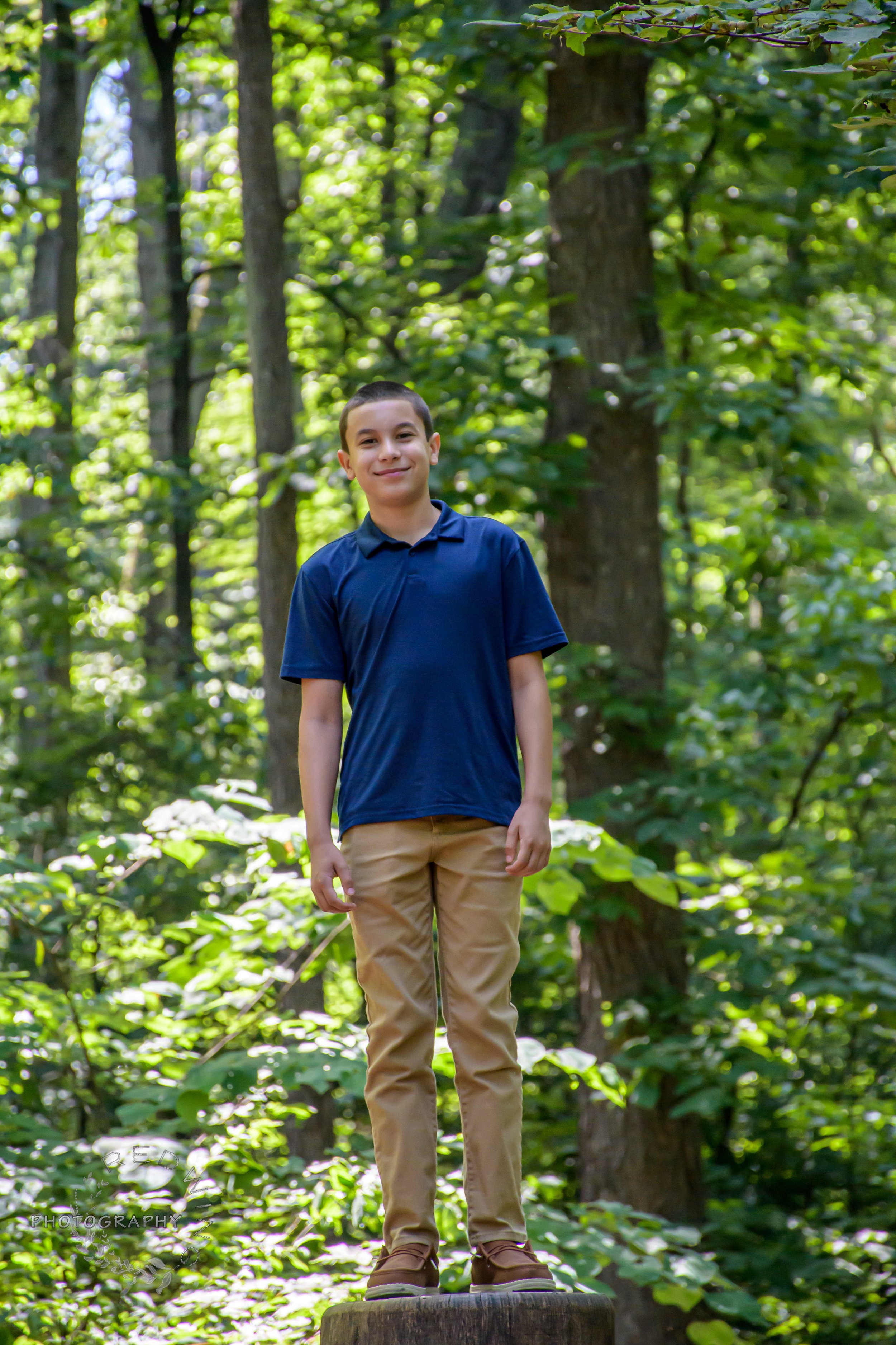A teenage boy with short dark hair wearing a navy blue polo shirt and tan pants standing on a tree stump in a lush green forest, smiling.