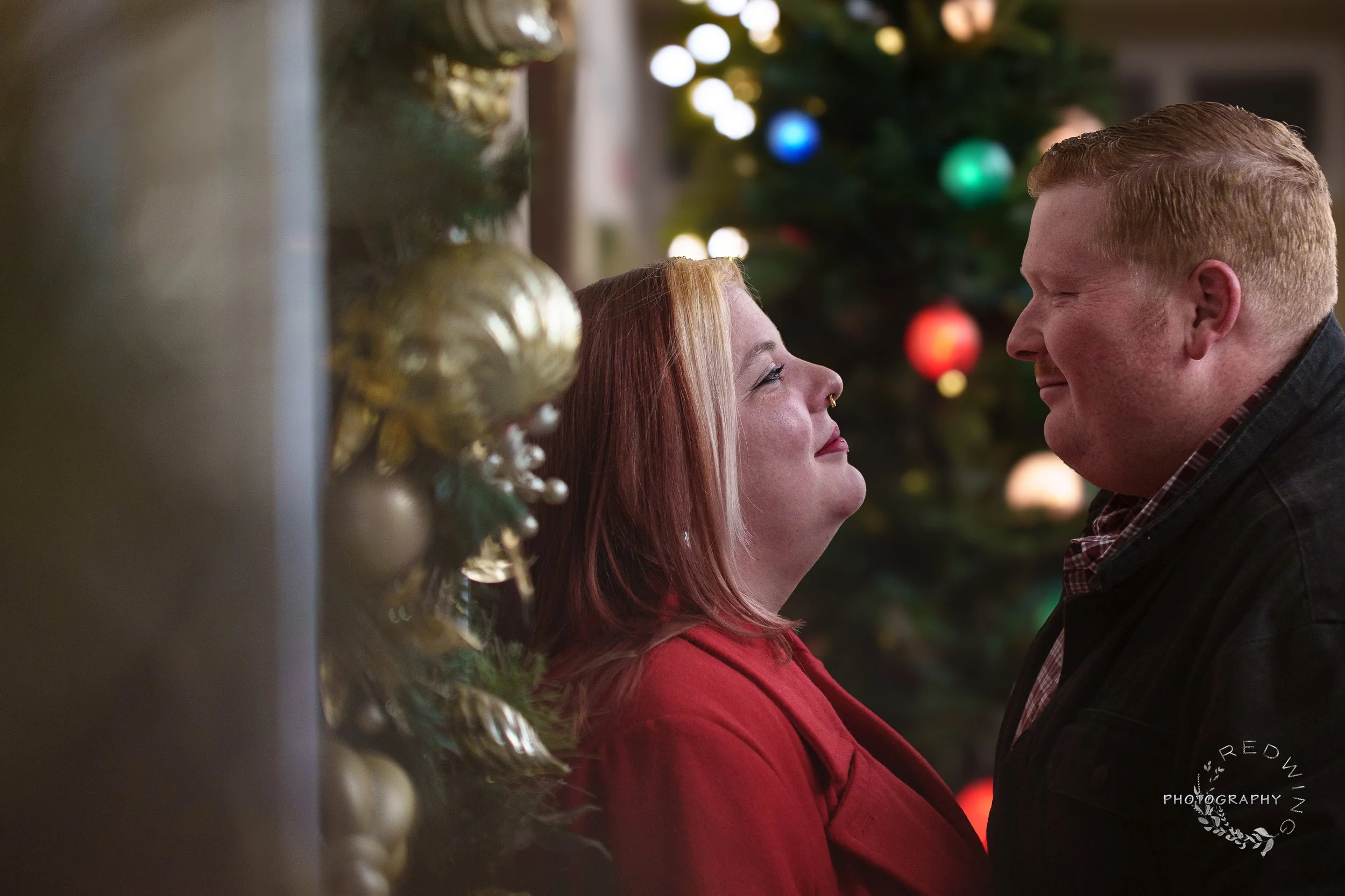 A couple standing close faces each other in front of a decorated Christmas tree, smiling with their foreheads touching.