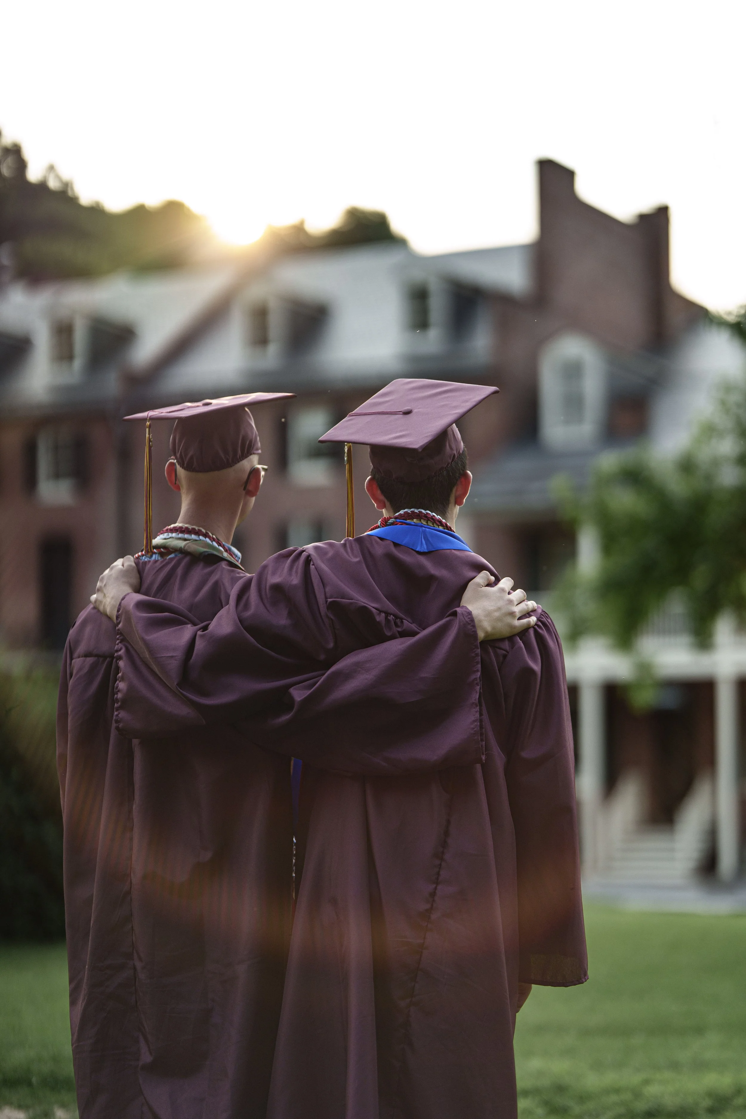 Two graduates in maroon caps and gowns with their arms around each other, celebrating outdoors during sunset in front of a residential building.