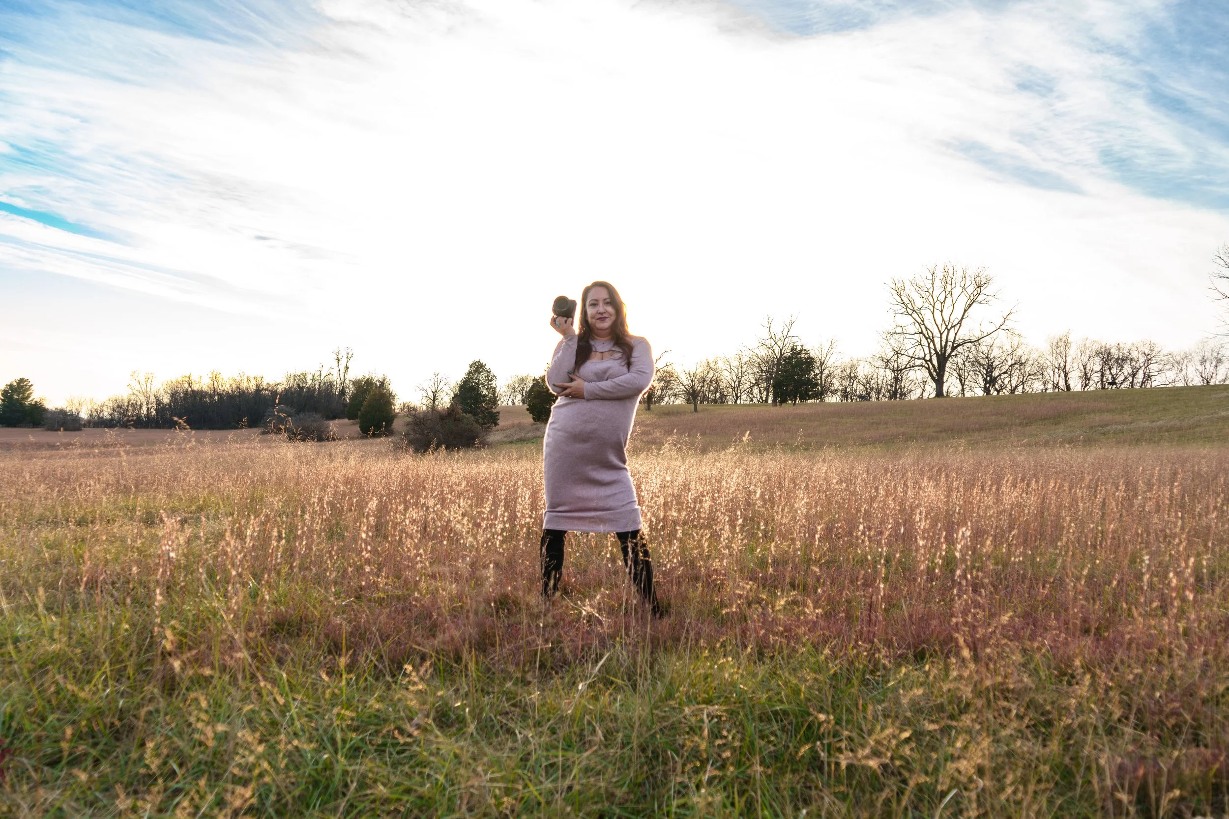 A woman standing in a field holding a DSLR camera, wearing a long, light-colored sweater dress with black patterned leggings, with a sunset sky and leafless trees in the background.