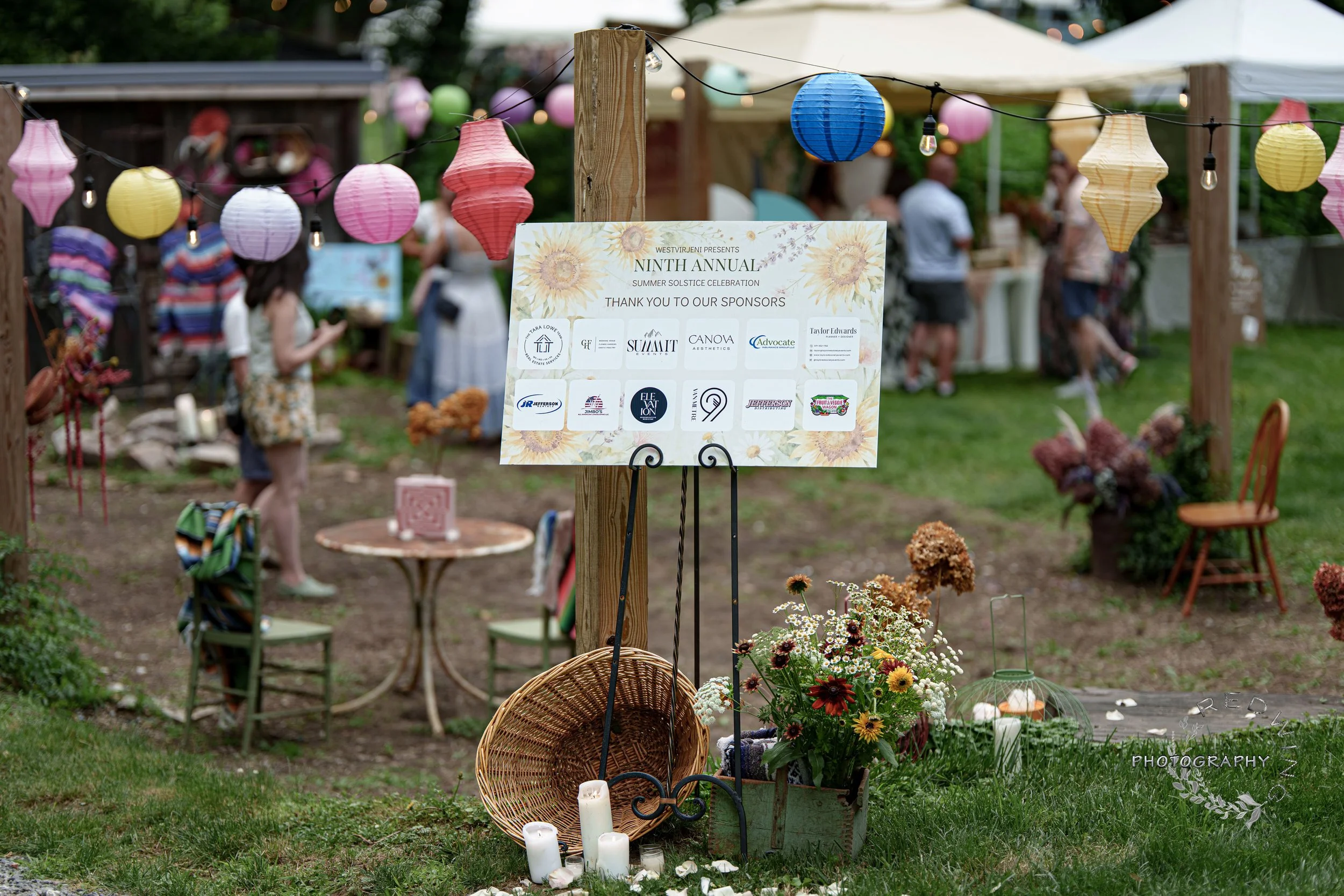 Memorial altar with candles, flowers, and a sign reading 'Ninth Annual Summer Solstice Celebration' at an outdoor gathering, decorated with colorful paper lanterns and string lights.