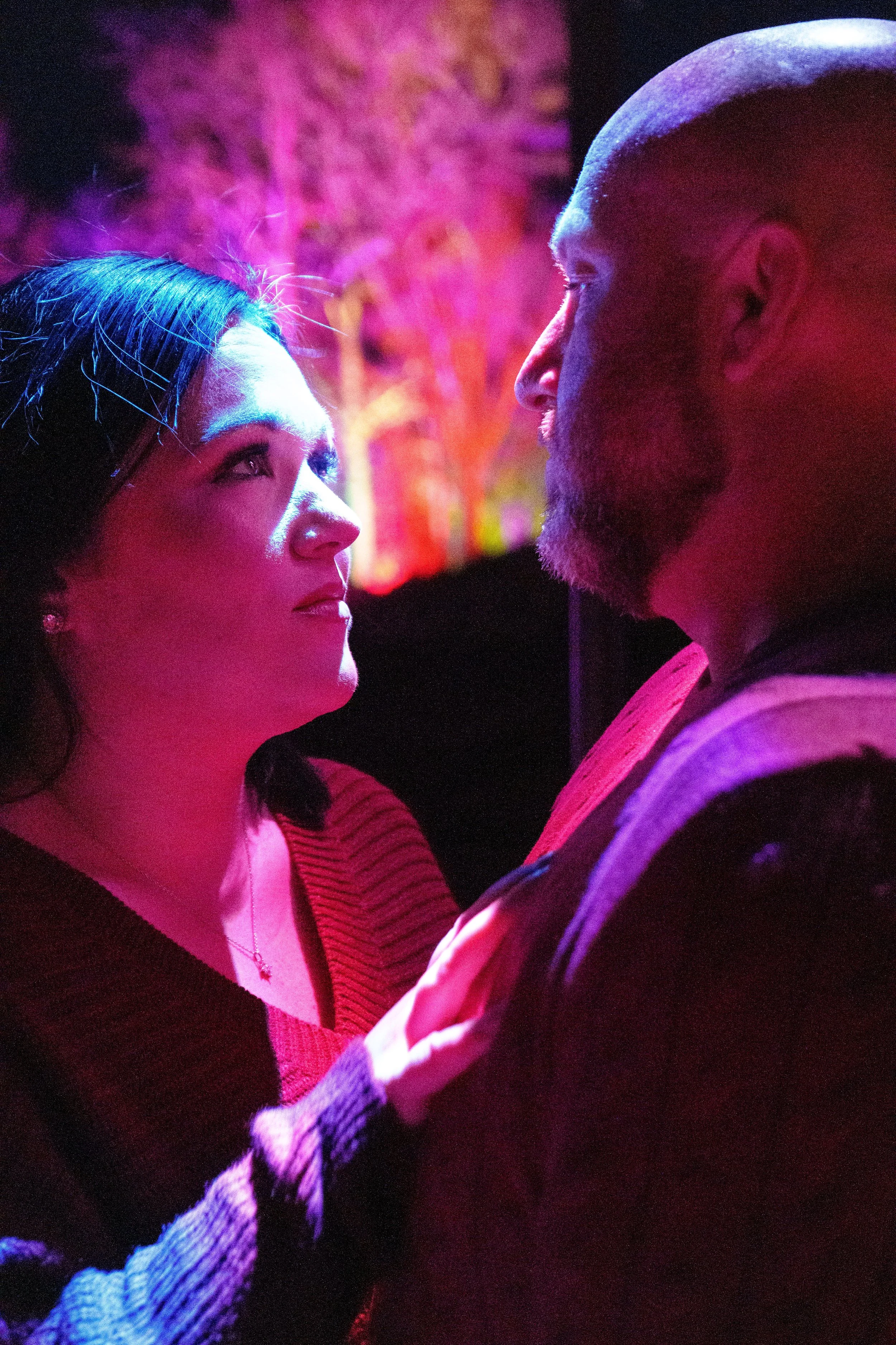 A woman and a man face each other in close-up, illuminated by vibrant pink, purple, and blue lighting, with a blurry colorful background.