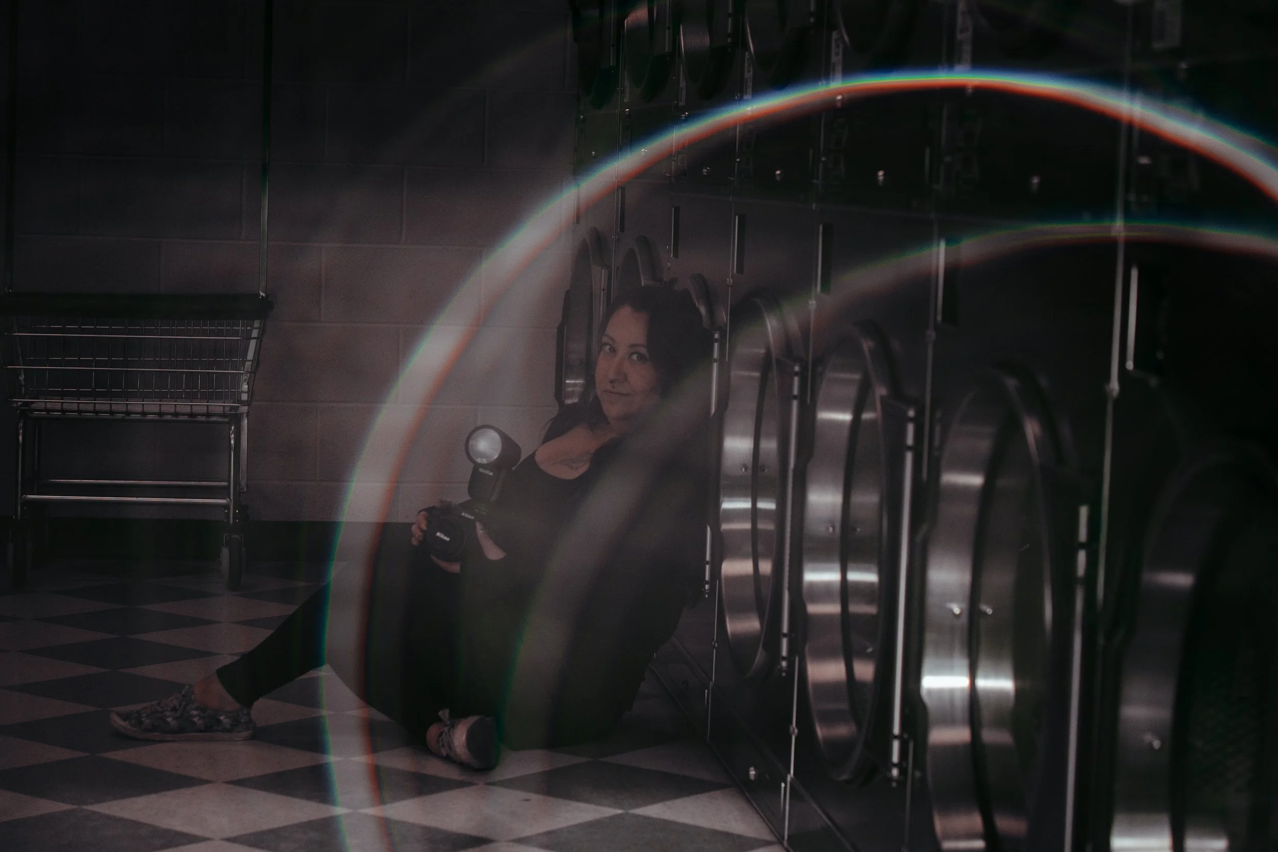 A woman sitting on the floor of a laundromat, holding a camera with a flash, beside a row of washing machines, with a rainbow-like lens flare across the image.