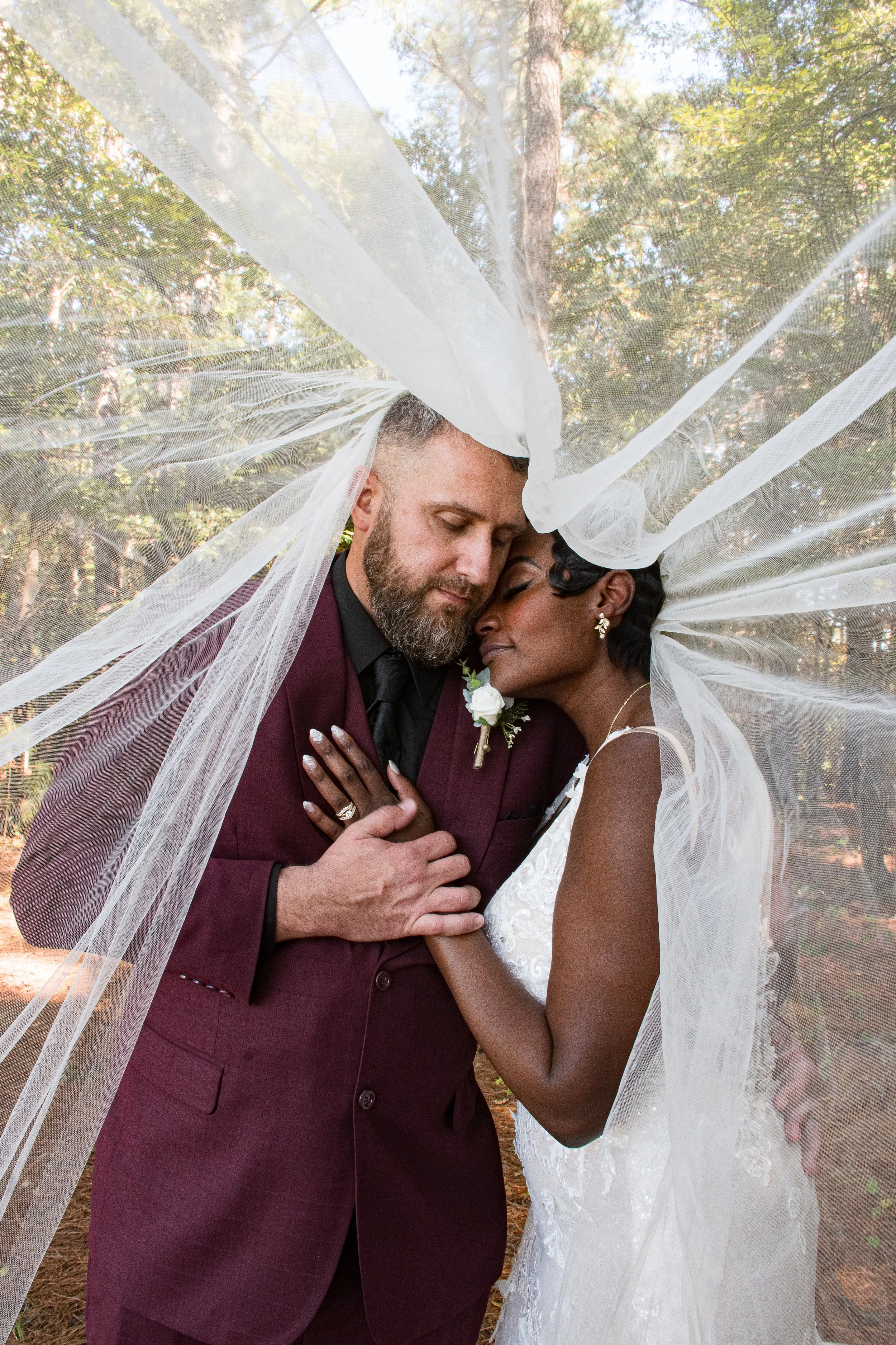 Couple on their wedding day, embracing outdoors surrounded by trees, with a sheer veil draped over their heads.