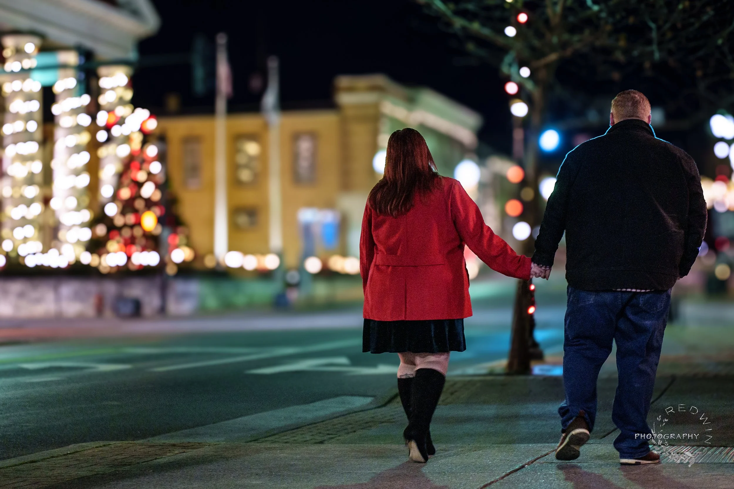 A couple holding hands and walking on a city sidewalk at night with blurred Christmas lights and buildings in the background.