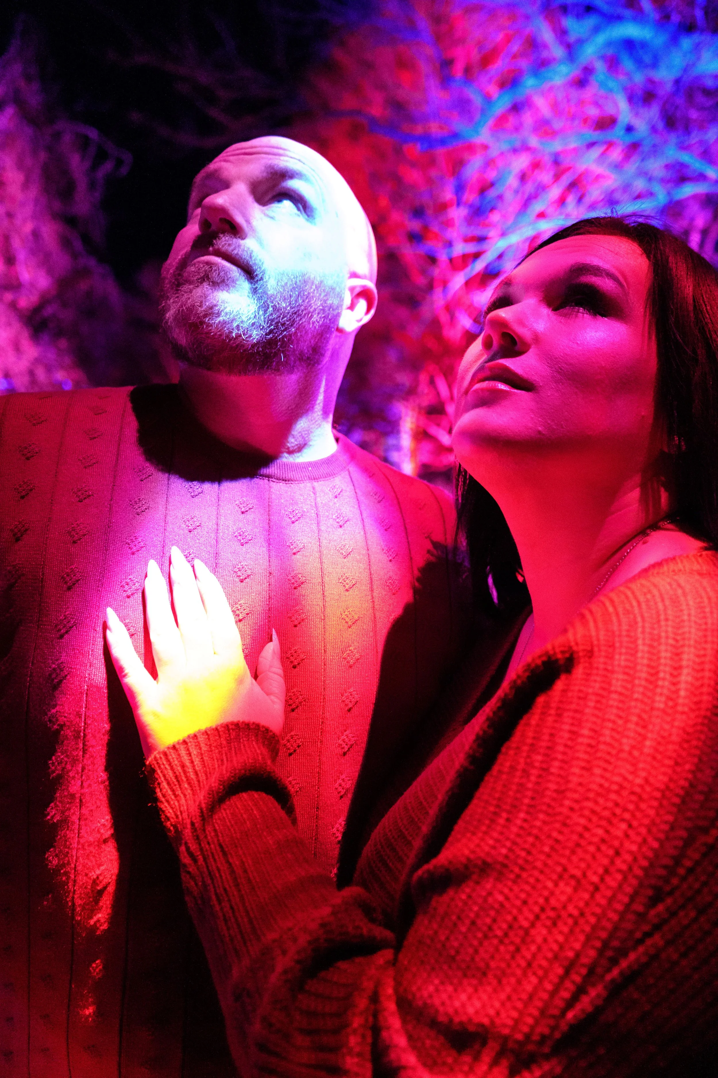 A man and woman pose under colorful, purple and pink lighting with a textured wall behind them. The woman is touching the man's chest with her hand, and both are looking upward.