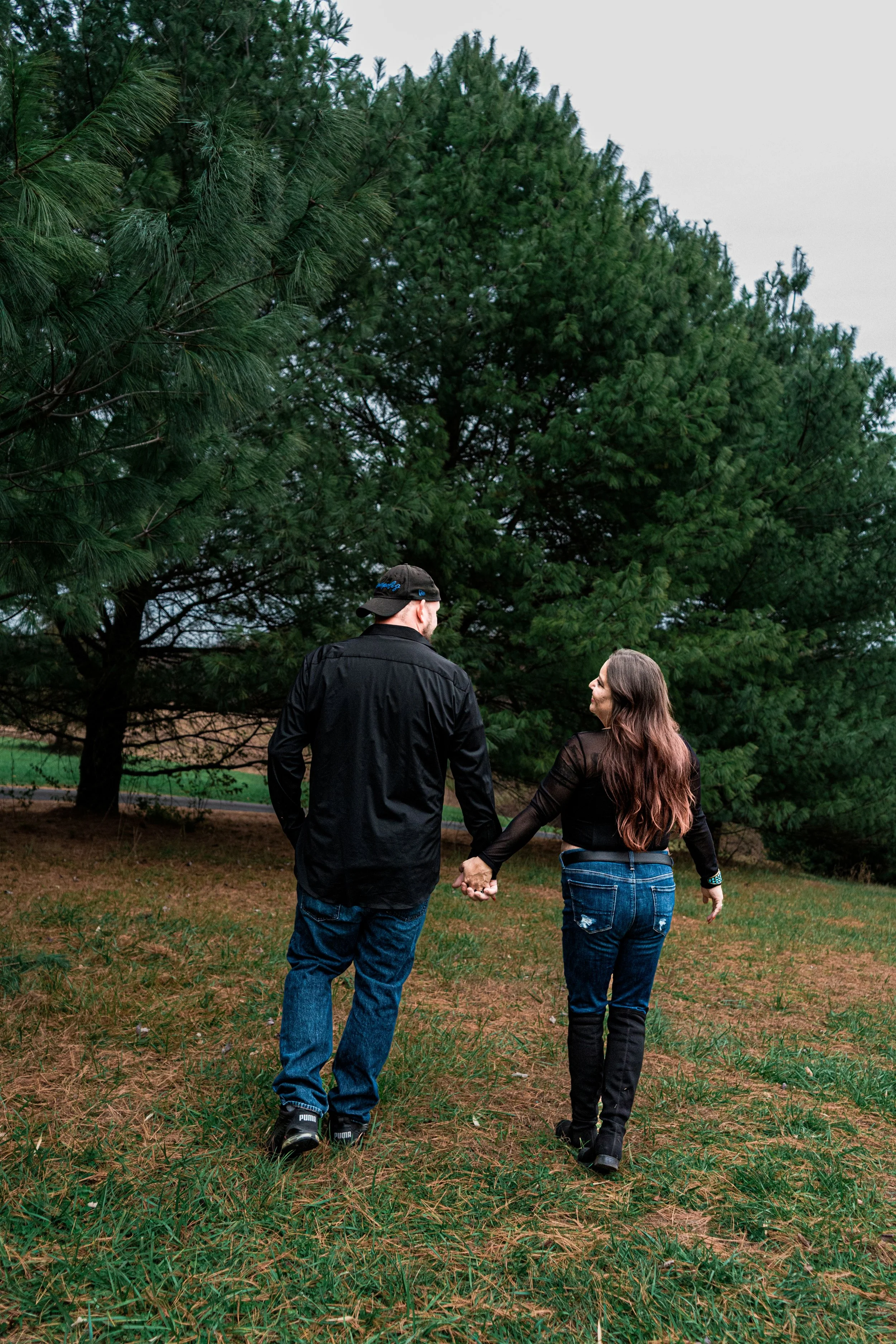 A couple holding hands walking through a park with tall green trees and grass.