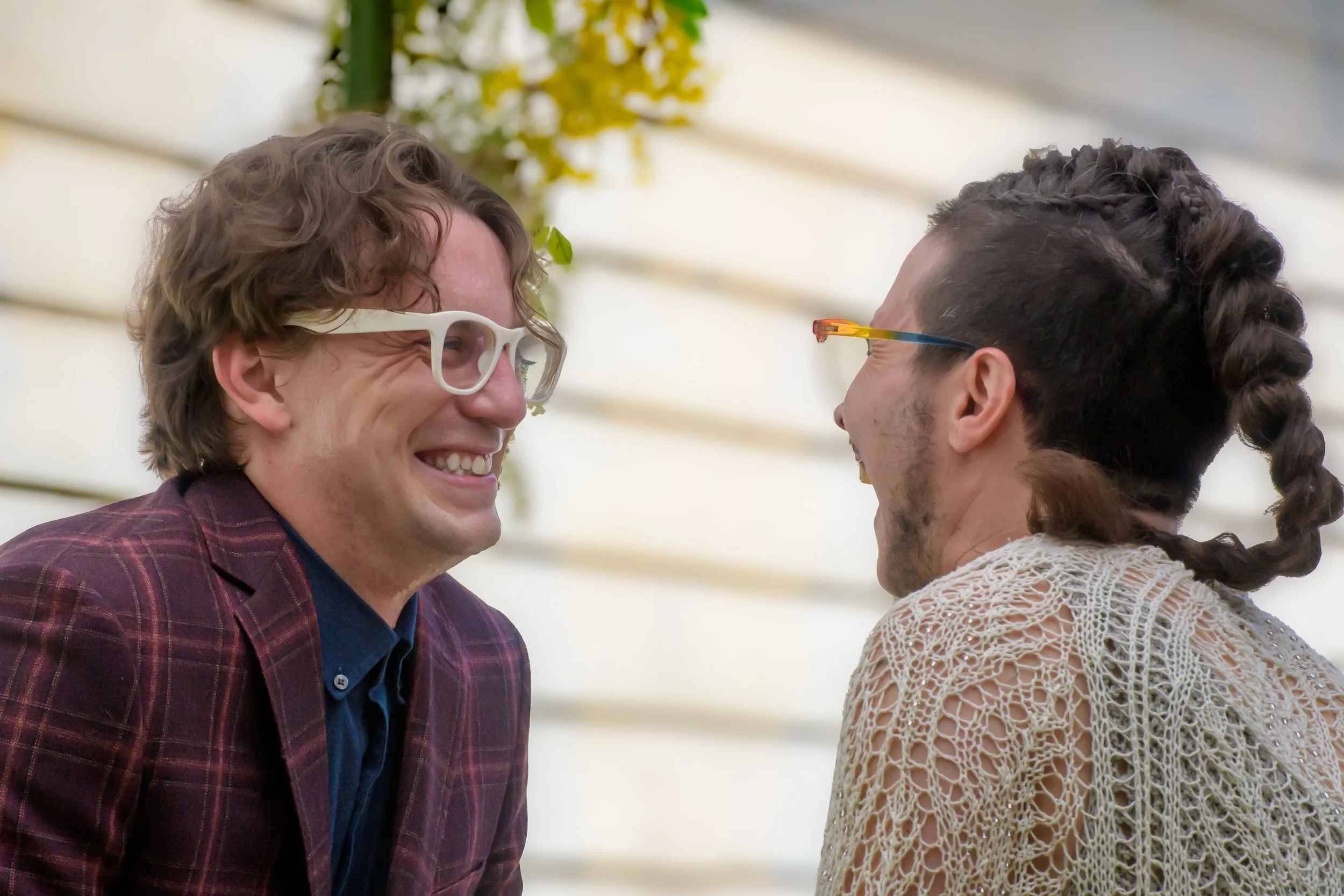 Two men are smiling and laughing while facing each other. One man has wavy hair, wears white glasses, and a plaid blazer. The other man has braided hair styled with curls, wears colorful glasses, and a crocheted top.