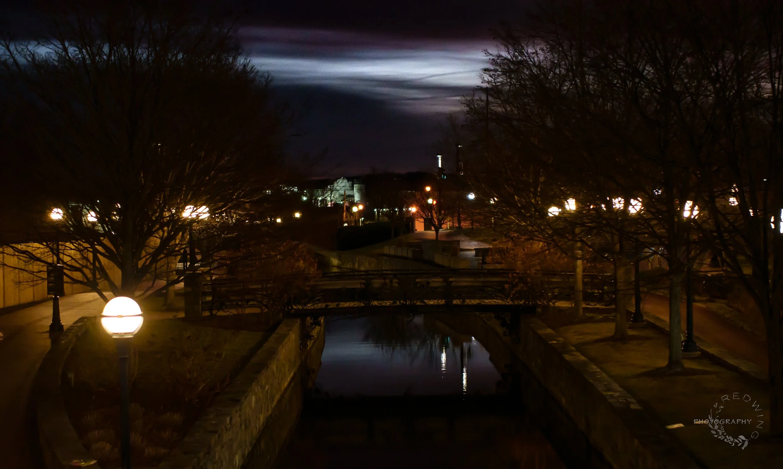 Nighttime scene of a park with a canal, lit by street lamps, with leafless trees lining the walkway and a bridge crossing over the canal, with buildings and streetlights in the background under a dark, cloudy sky.