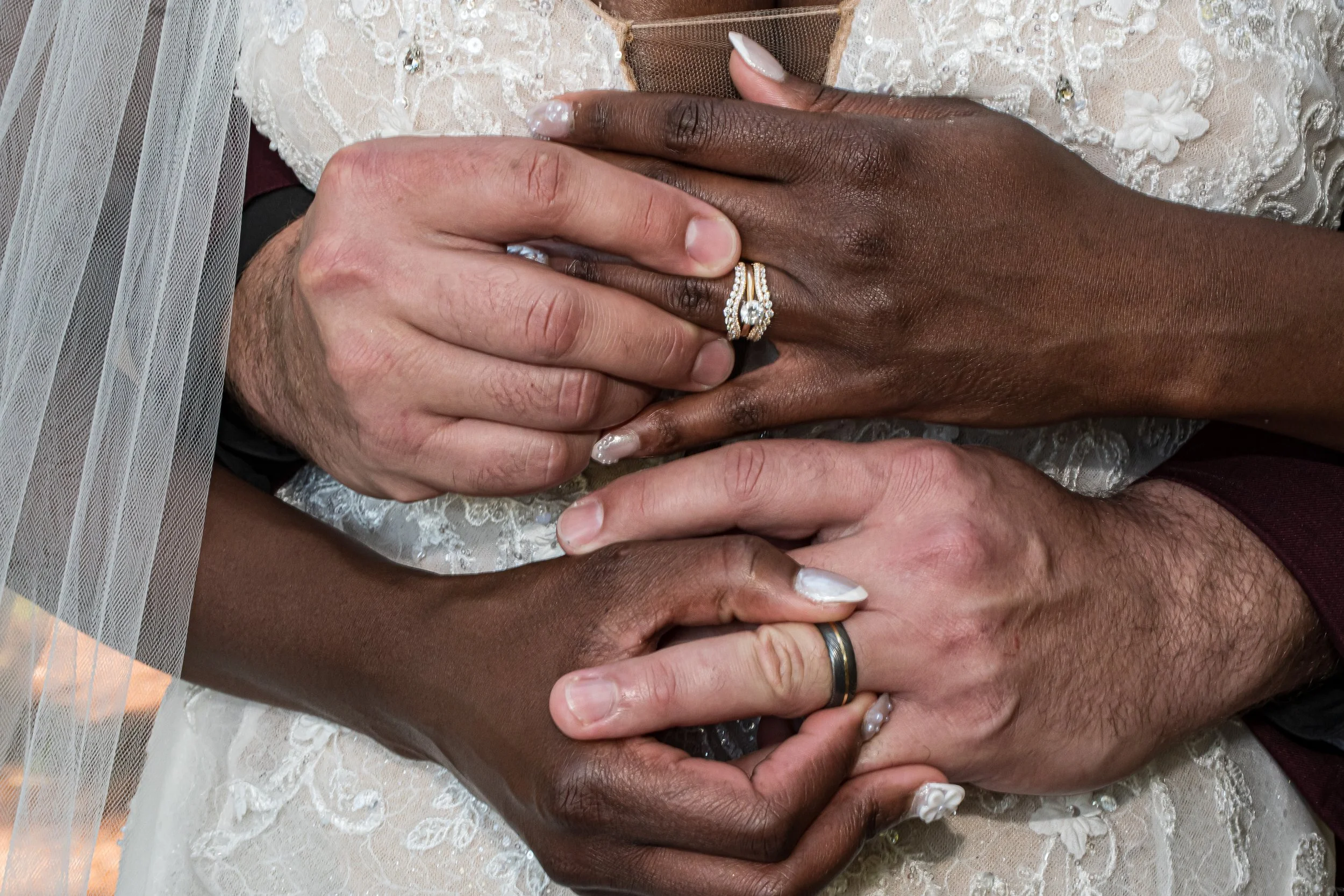 A close-up of three hands, two with darker skin and one with lighter skin, joined together in a gesture of unity. The darker-skinned individual is wearing a wedding band and an engagement ring, while the lighter-skinned person has a wedding band. The background shows part of a wedding dress and veil.