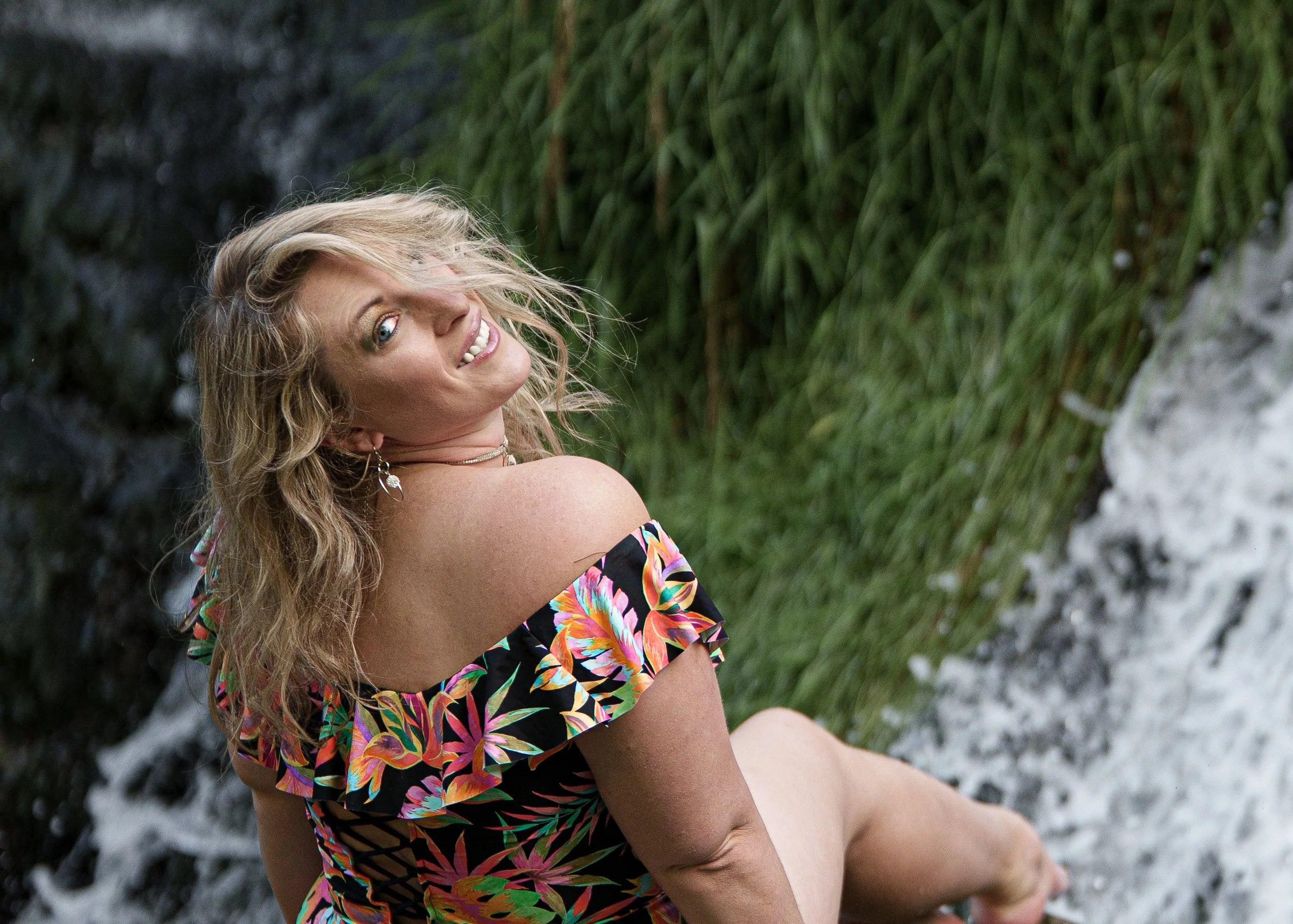 A woman with curly blonde hair, wearing earrings and a colorful, tropical-patterned off-the-shoulder dress, sitting outdoors near greenery and rocks, smiling at the camera.