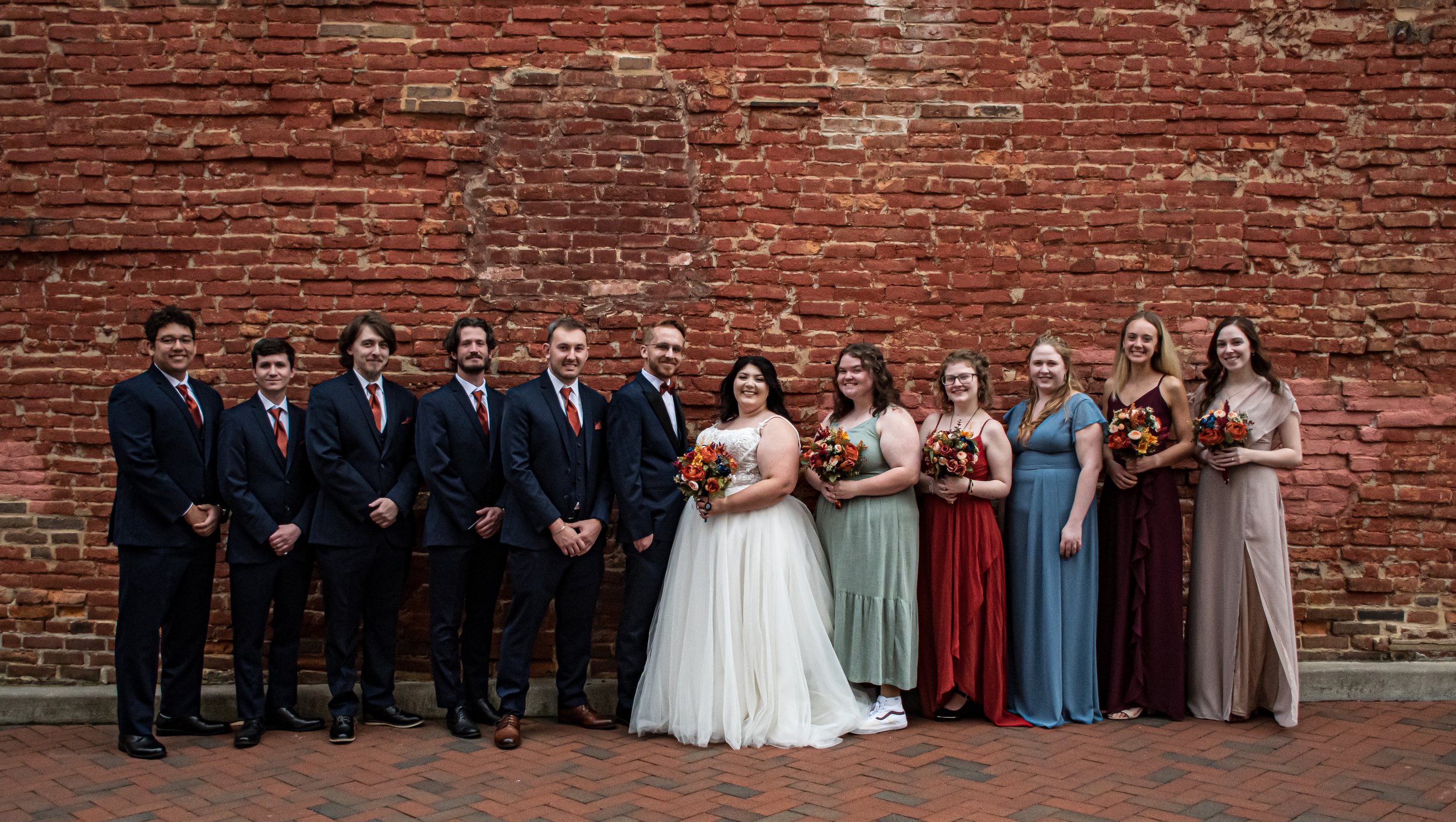 A group of eleven people, wearing formal attire, standing against a red brick wall. Four men dressed in dark suits and ties, and seven women in colorful dresses holding bouquets of flowers, celebrating a wedding.
