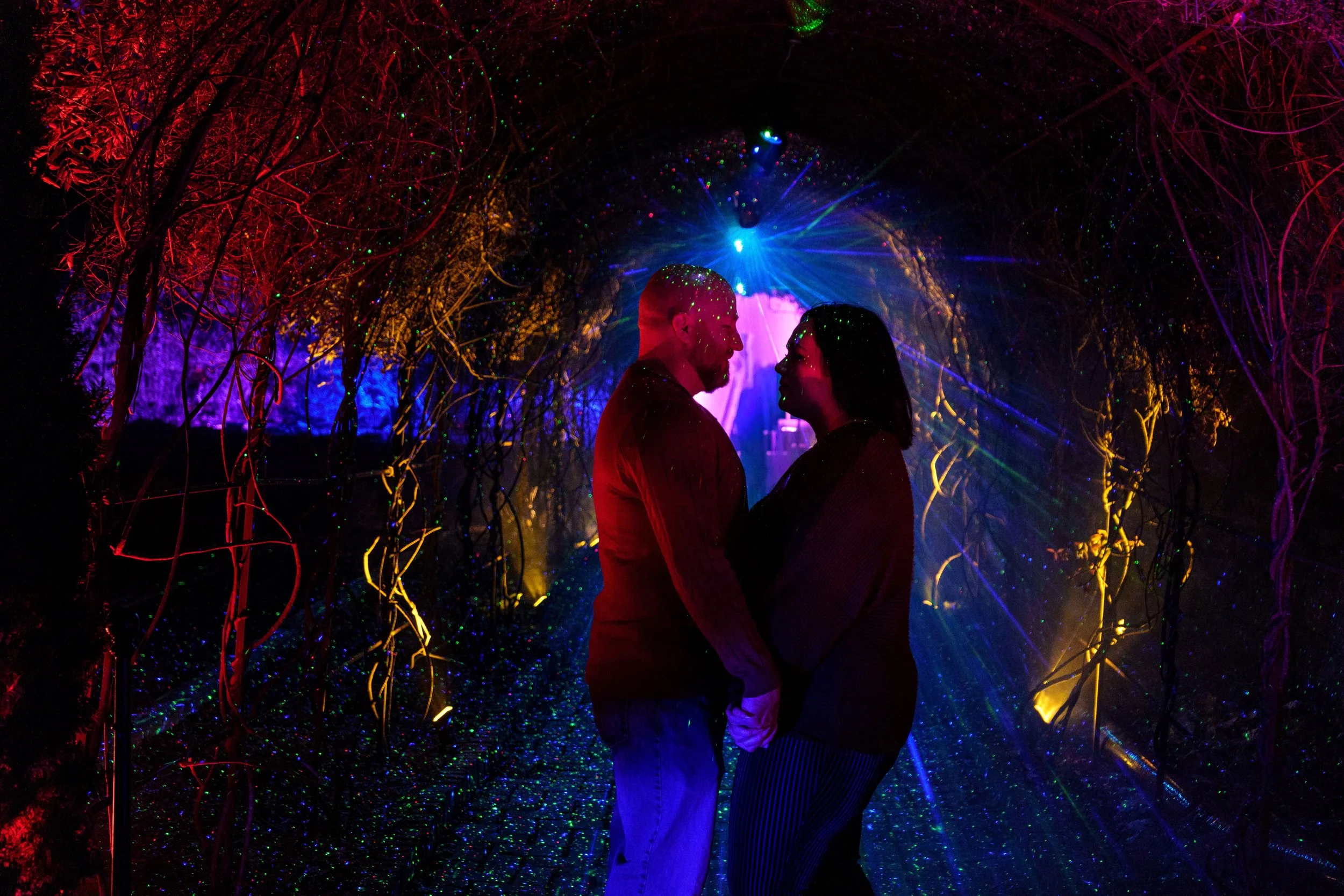 Couple holding hands and facing each other in a colorful, illuminated tunnel with vibrant, glowing lights and intricate vine-like structures.