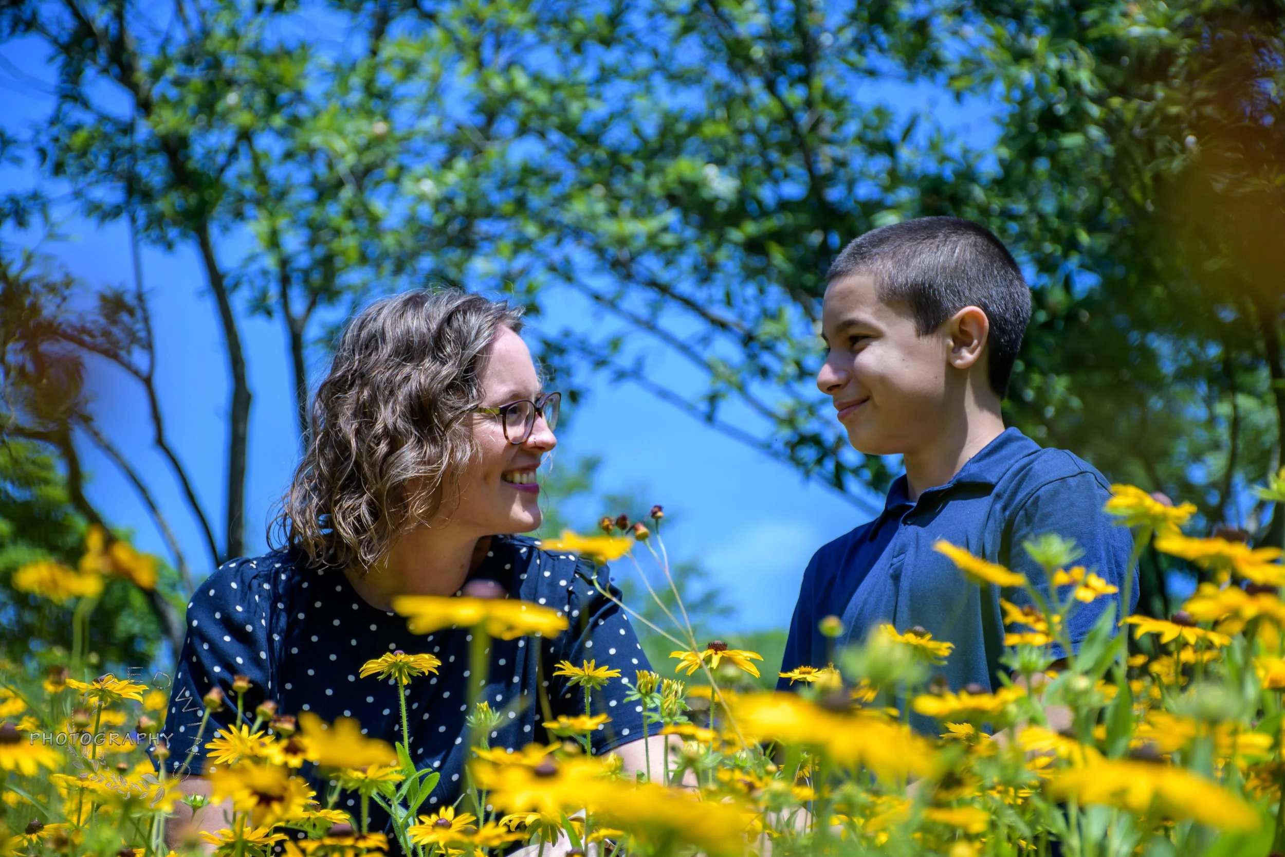 A woman and a boy smiling at each other among yellow flowers outdoors on a sunny day.