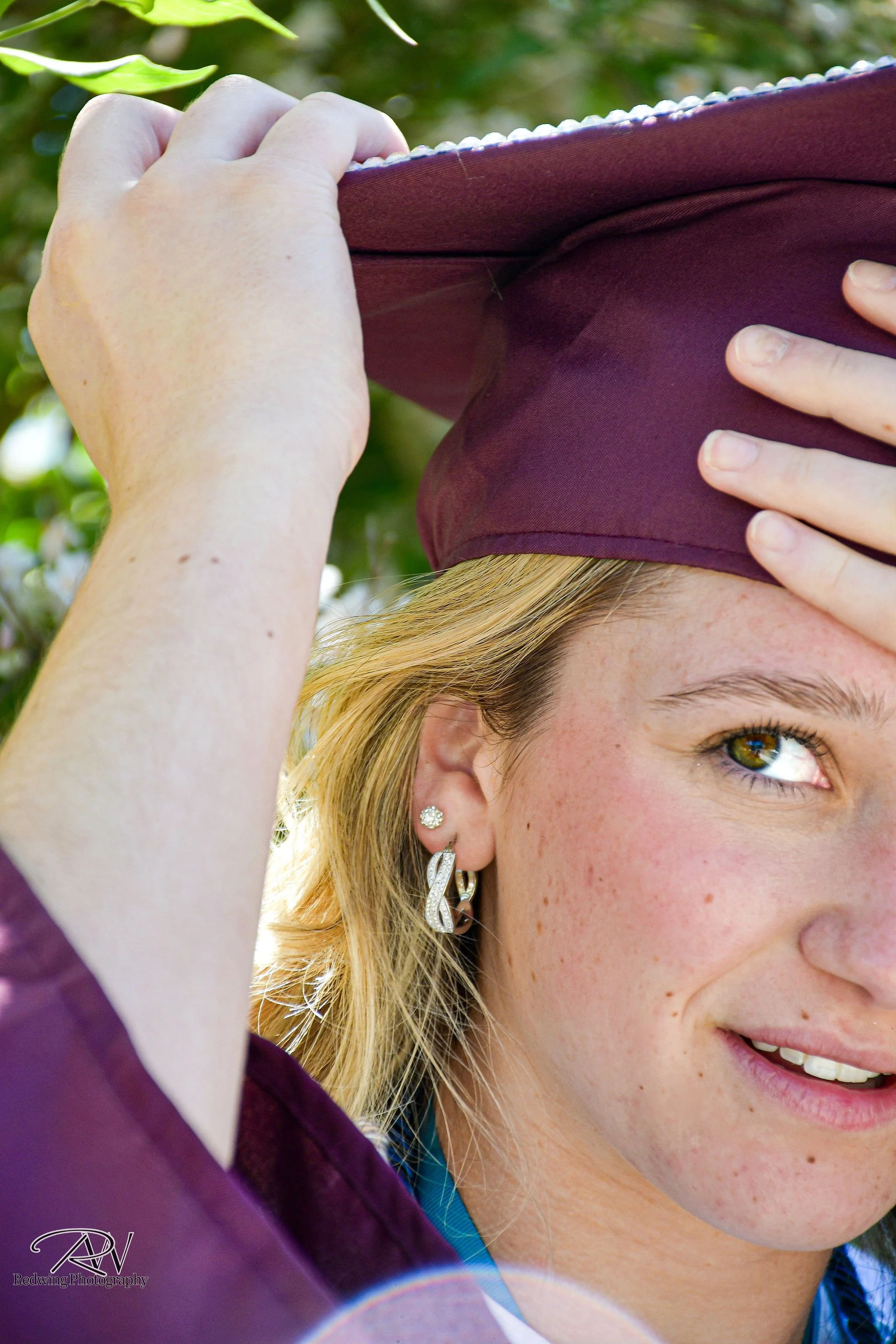 A young woman with blonde hair and earrings is smiling, wearing a maroon graduation cap and gown, holding her cap with one hand and looking to the side.