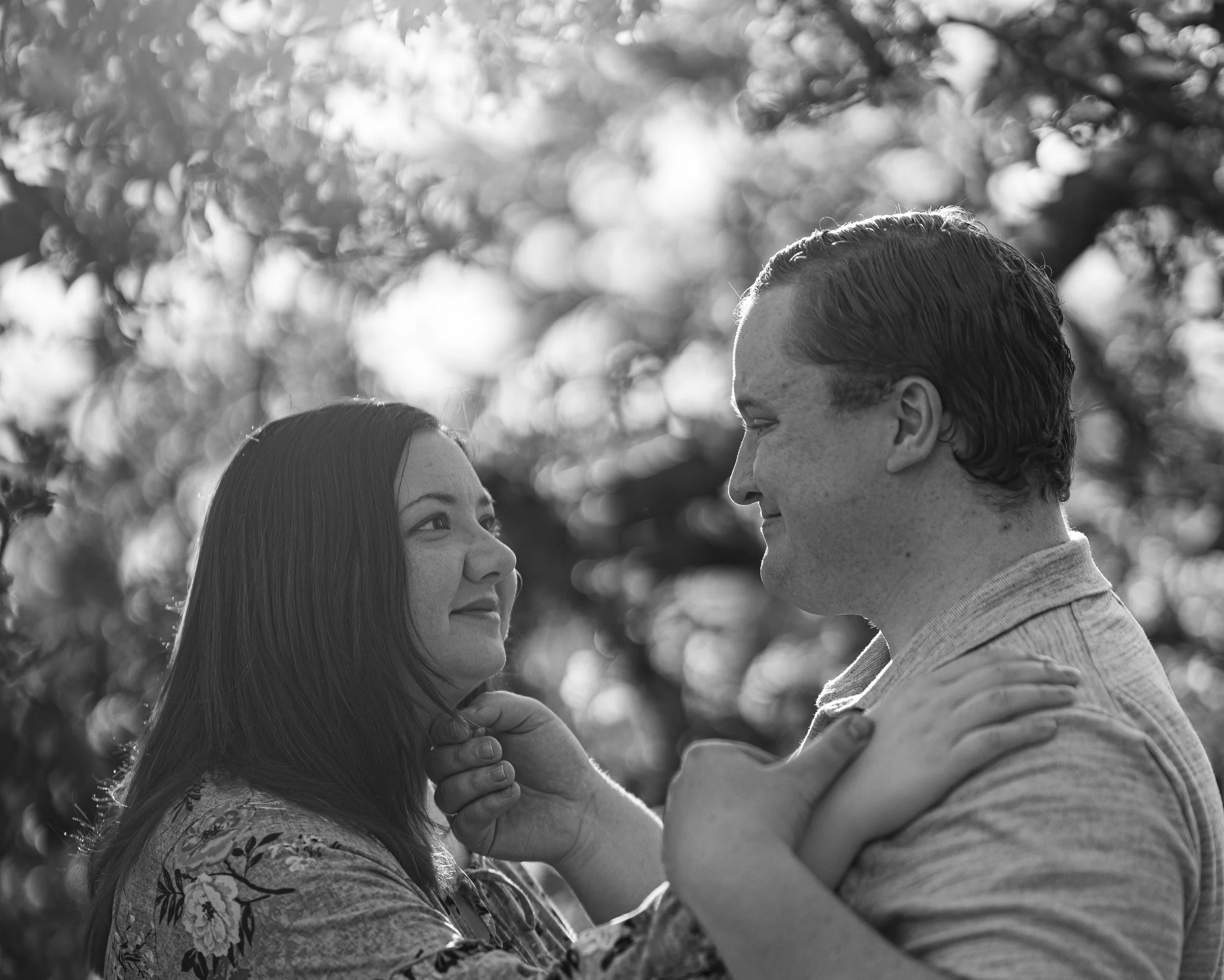 A black and white photograph of a man and woman gazing into each other's eyes outdoors surrounded by trees.