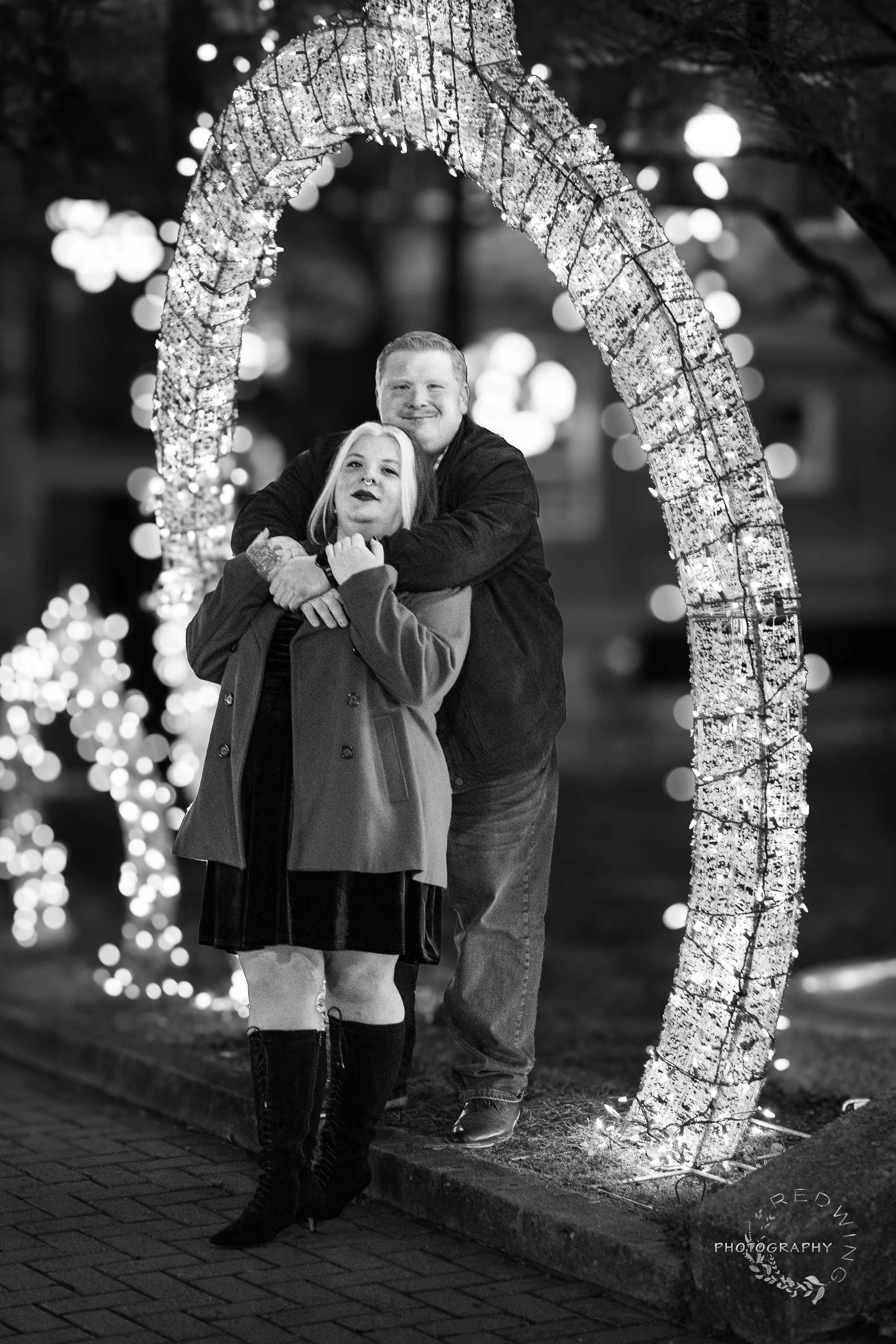 A black and white photo of a couple standing under a lit decorative arch at night, with festive lights in the background.