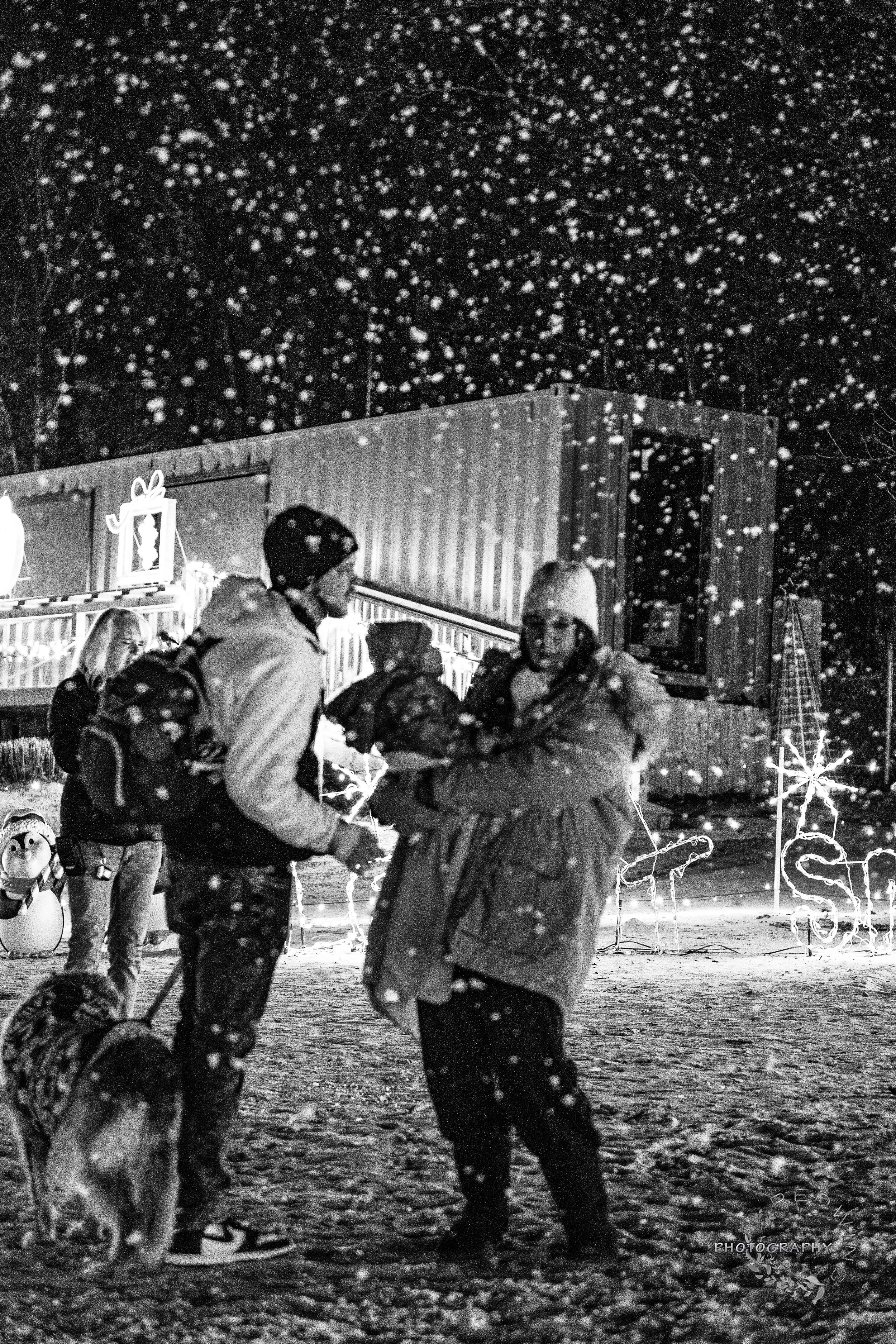 People exchanging gifts outside in the snow at night, decorated with holiday lights and Christmas decorations.