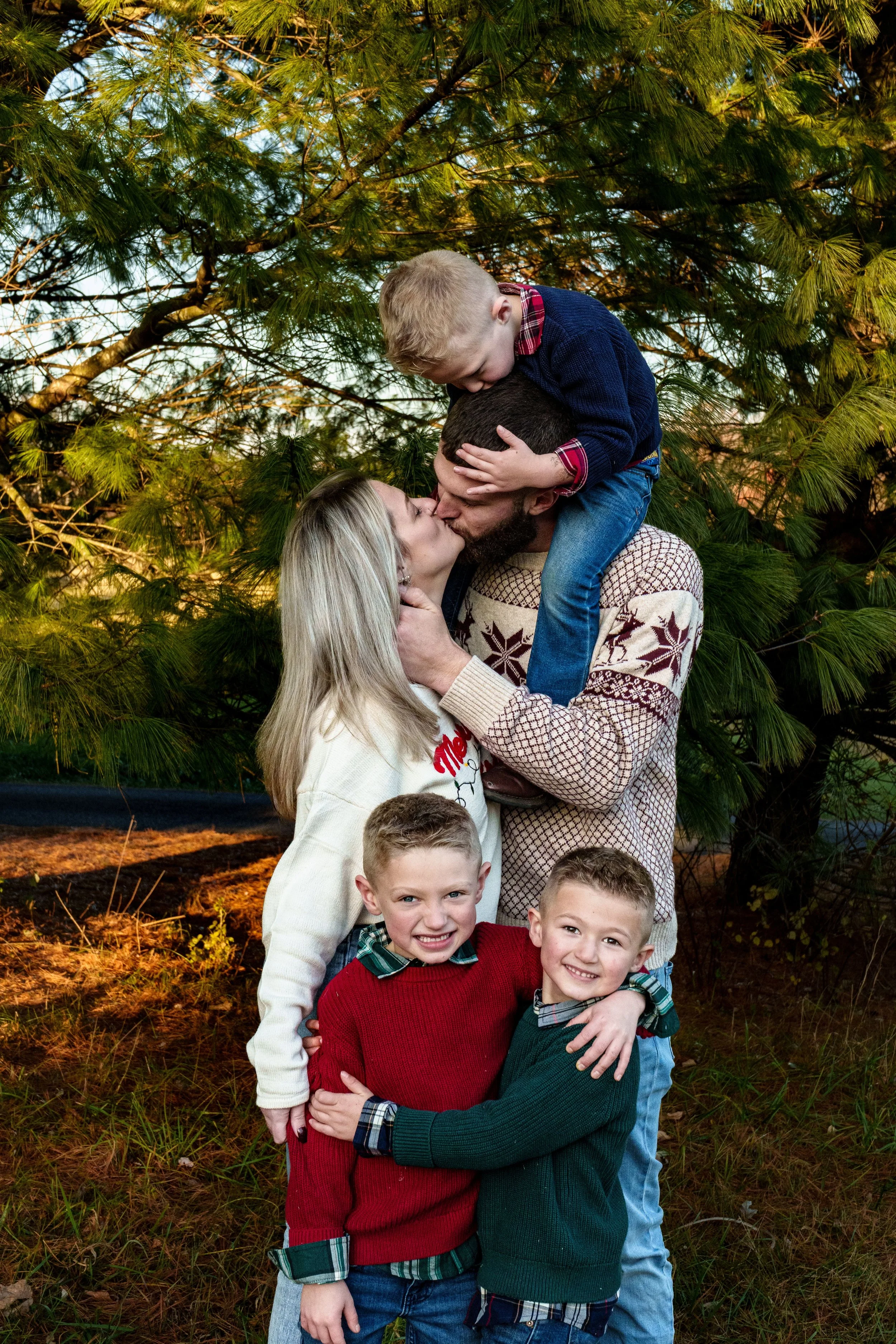 A family of five sharing a heartfelt moment outdoors during daytime, surrounded by green pine trees, with parents kissing and the children embracing each other.