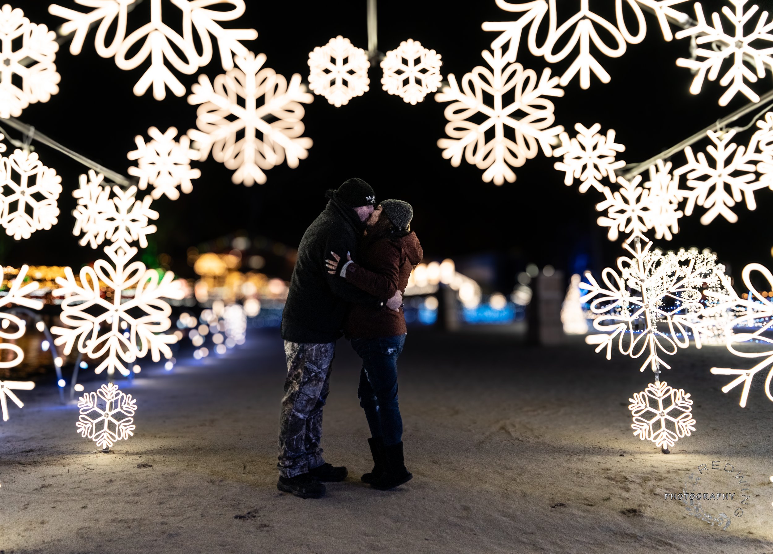 A couple sharing a kiss under large illuminated snowflake decorations at night, with colorful blurred lights in the background.
