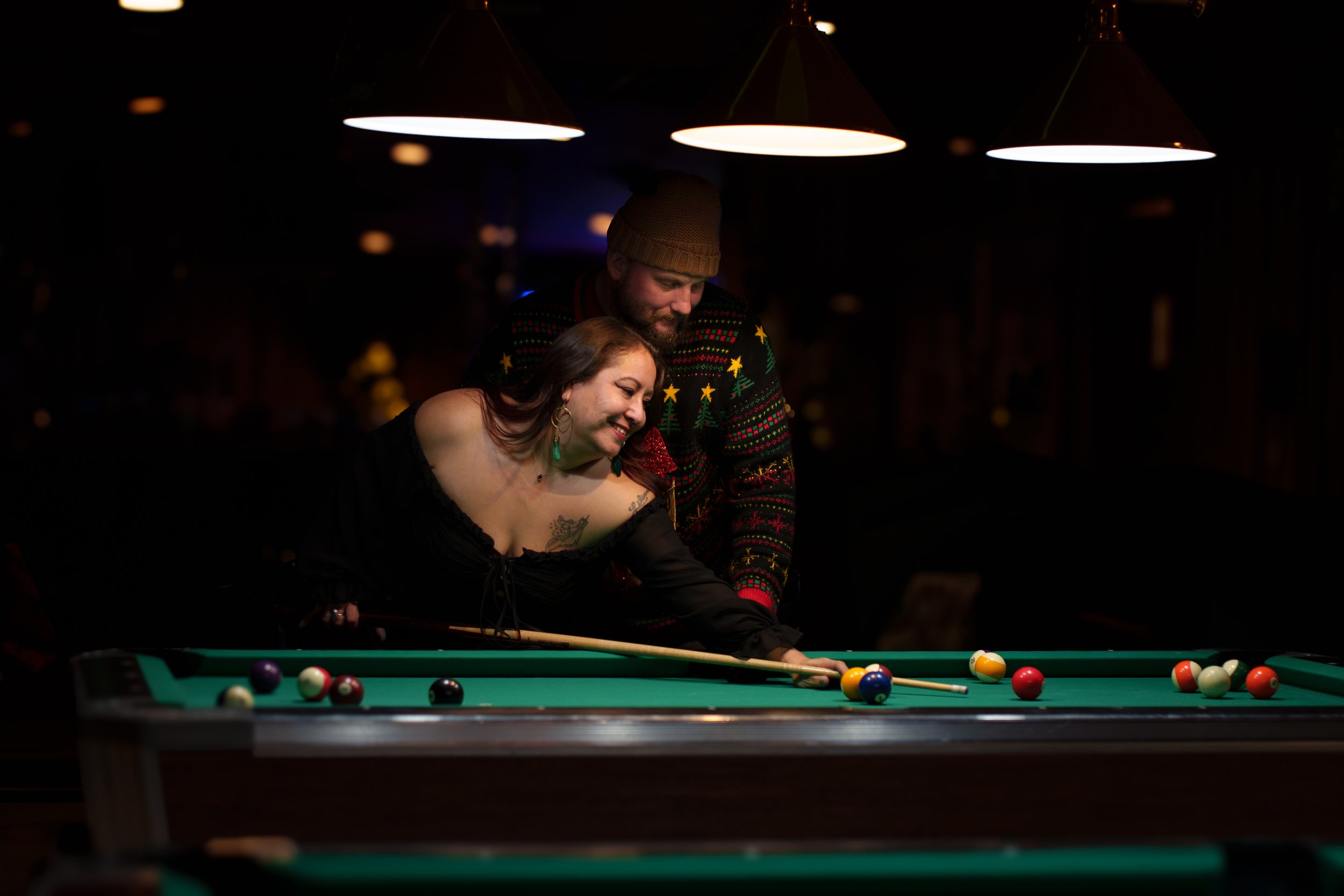 A woman and man playing pool at a dark bar or pool hall, with overhead lights illuminating the table. The woman is taking a shot, leaning over the table, and smiling, while the man stands behind her, watching closely, both appearing happy and engaged.