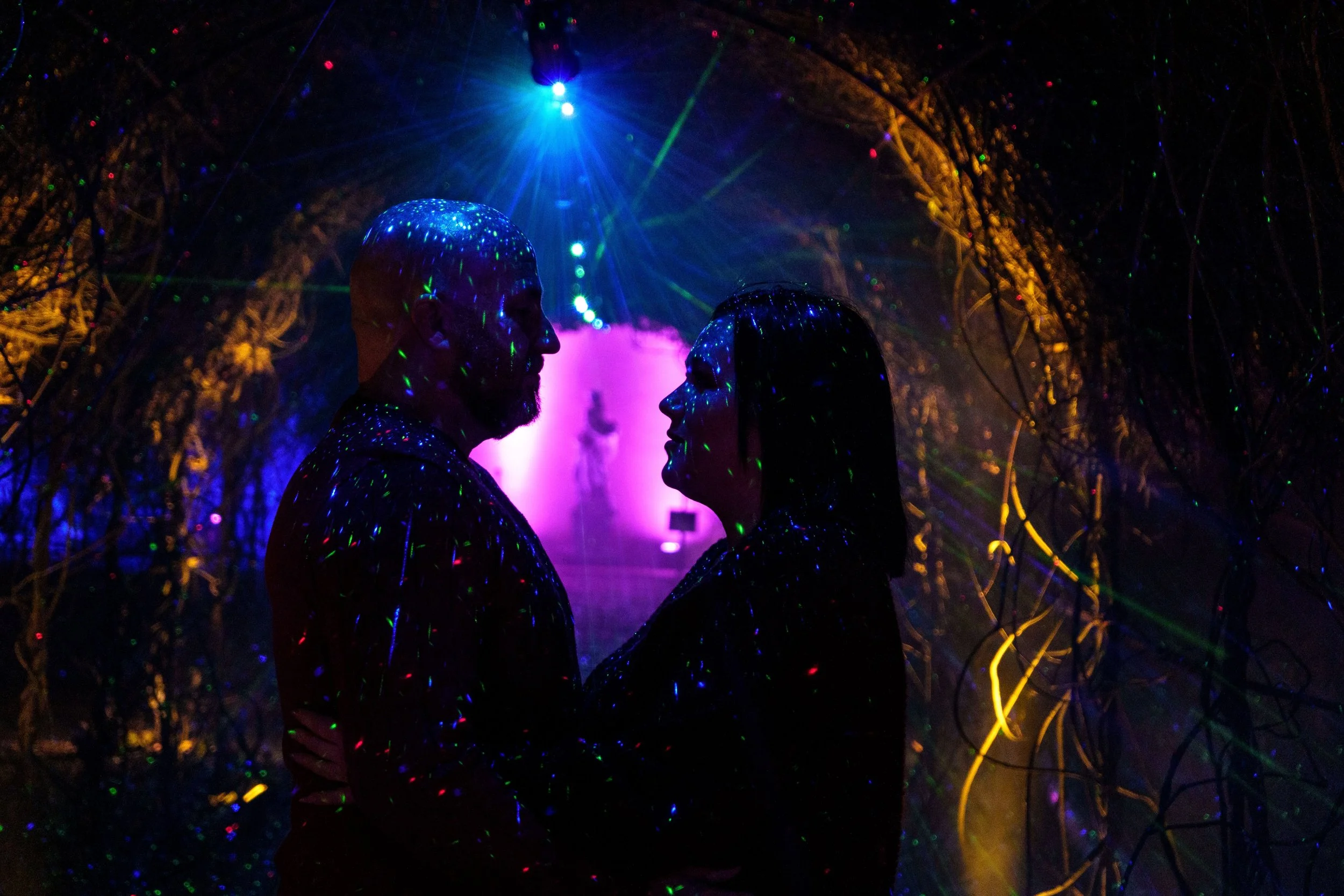 Two people standing close and facing each other in a dark, tunnel-like space illuminated by colorful lights, with a purple light and a bright blue light above.