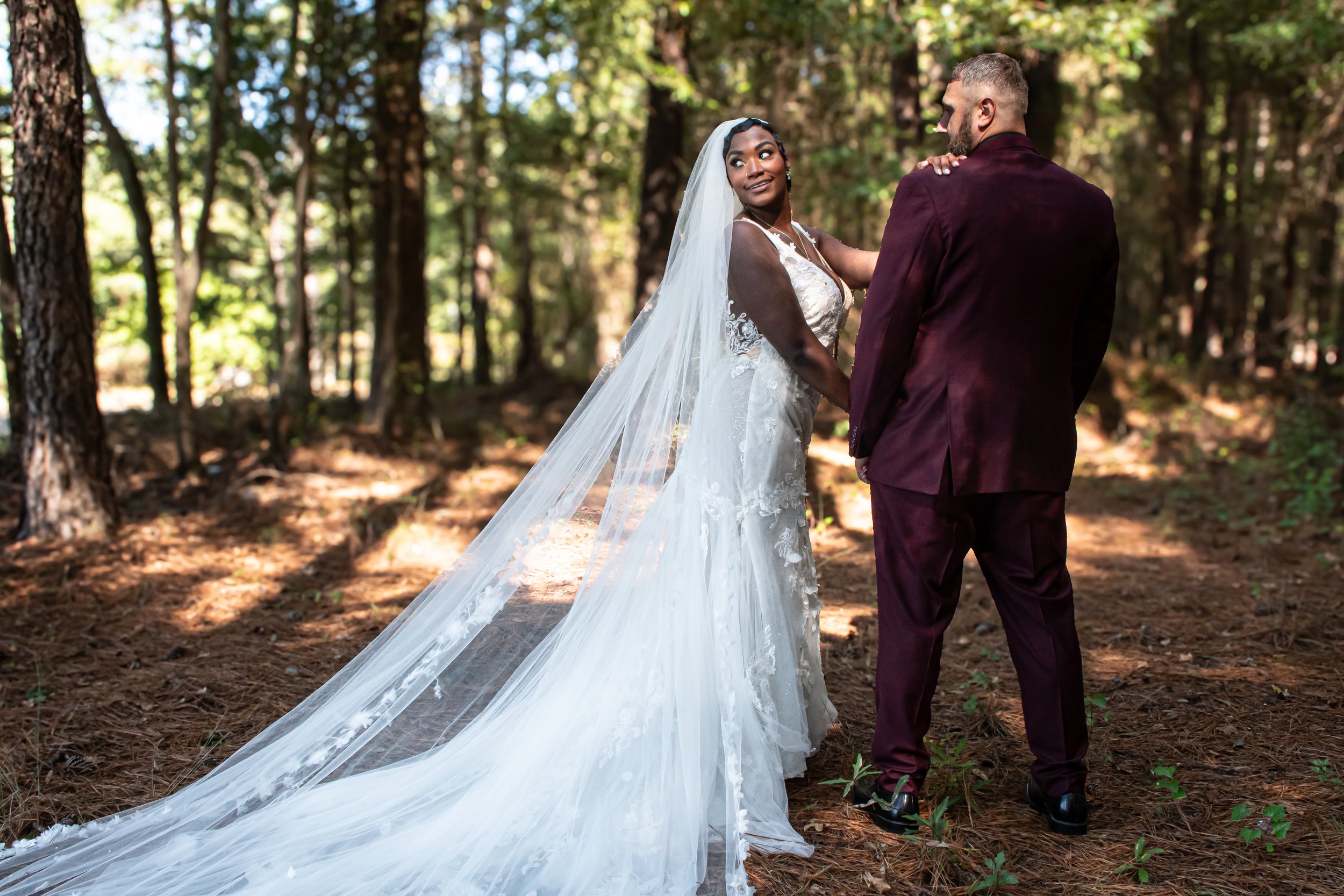 A bride and groom in formal attire standing in a wooded forest, the bride looking at the camera and the groom looking at her.