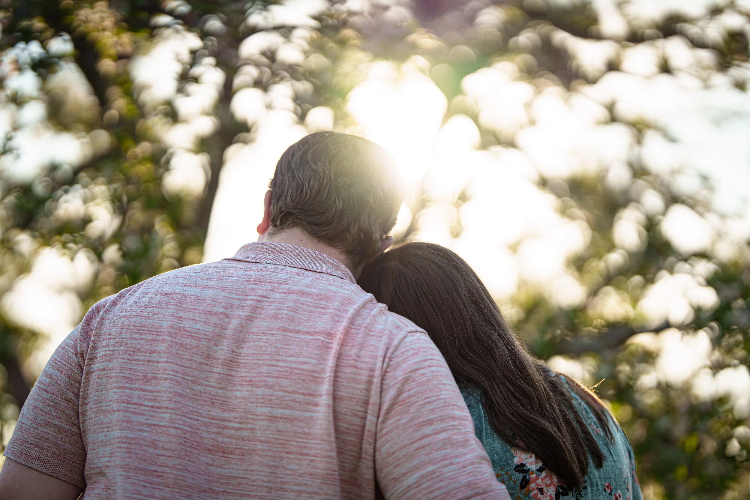 A couple with their heads close together outdoors with sunlight shining behind trees.