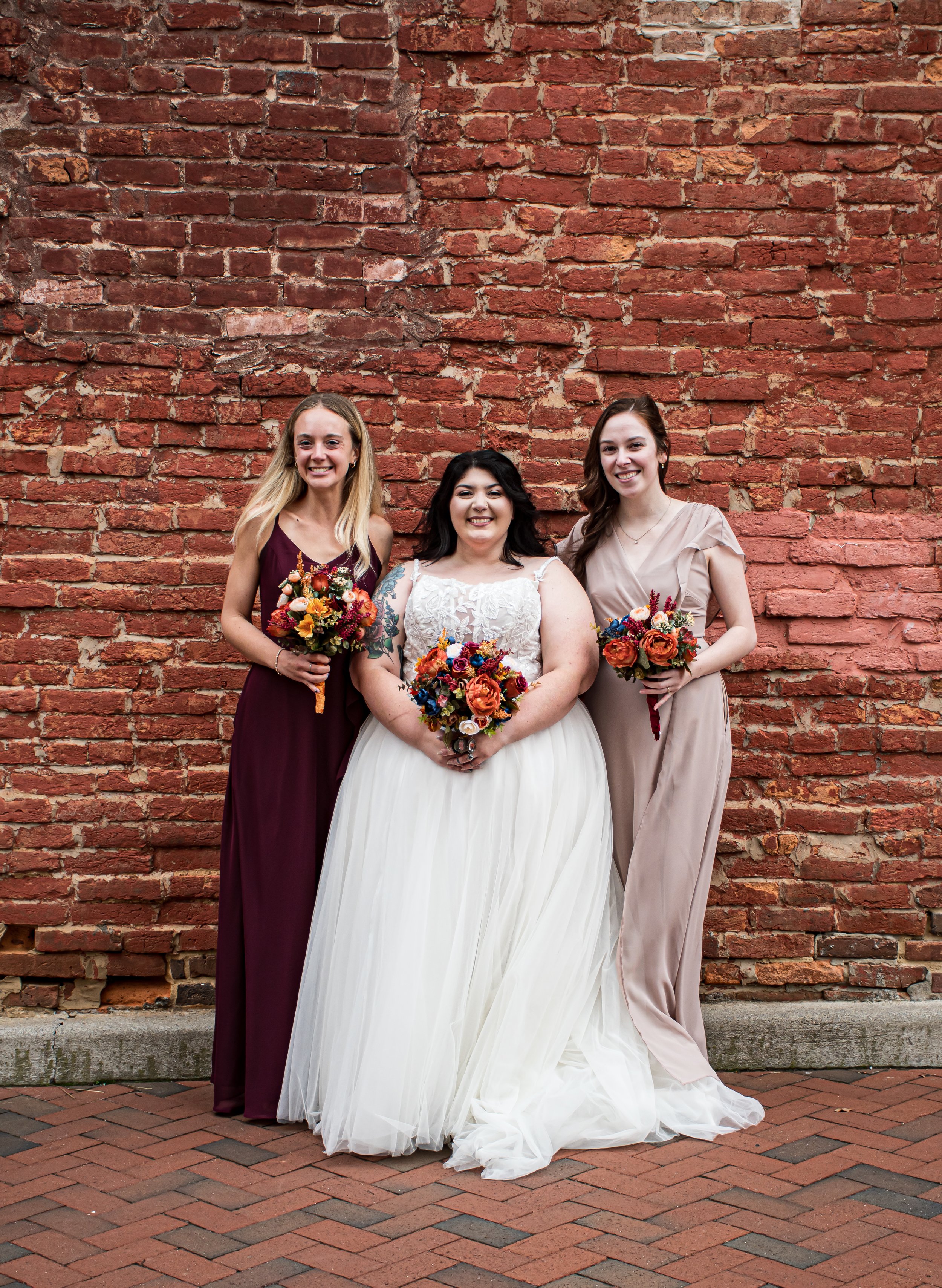 A bride and two bridesmaids standing against a red brick wall, smiling, holding bouquets of colorful flowers.