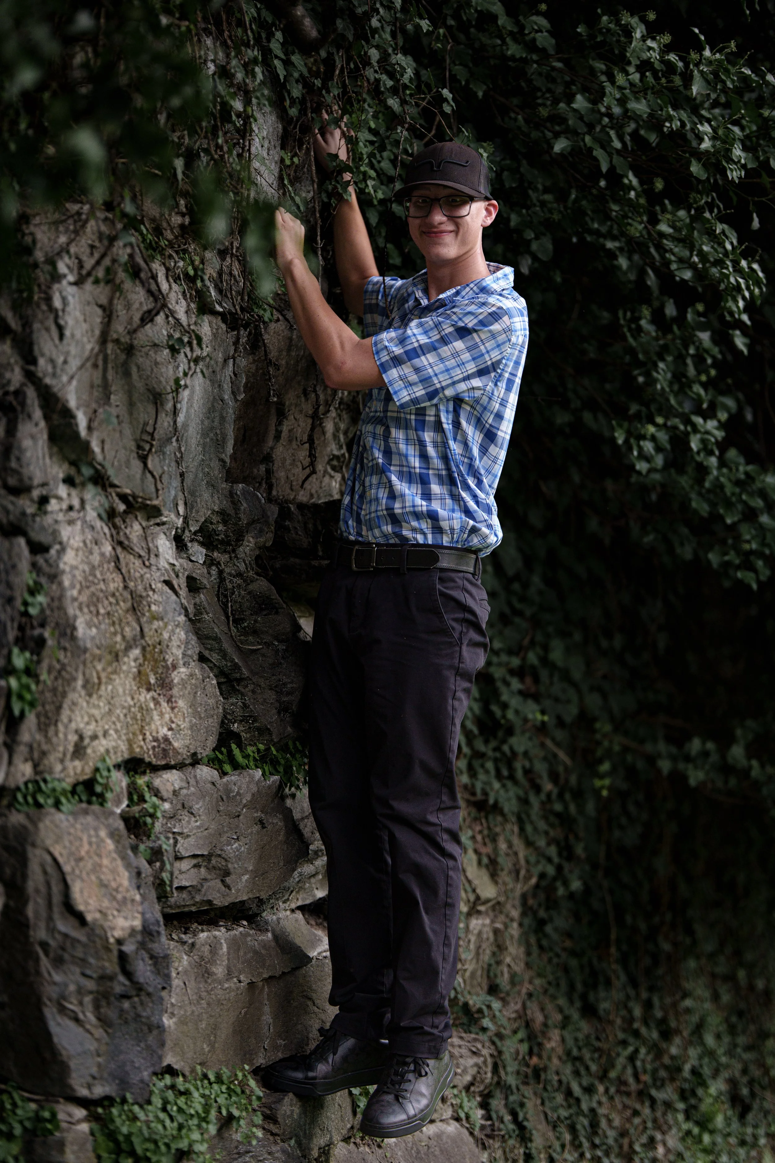 Young man wearing glasses, a baseball cap, a blue plaid shirt, and black pants climbing a stone wall surrounded by green ivy and foliage.