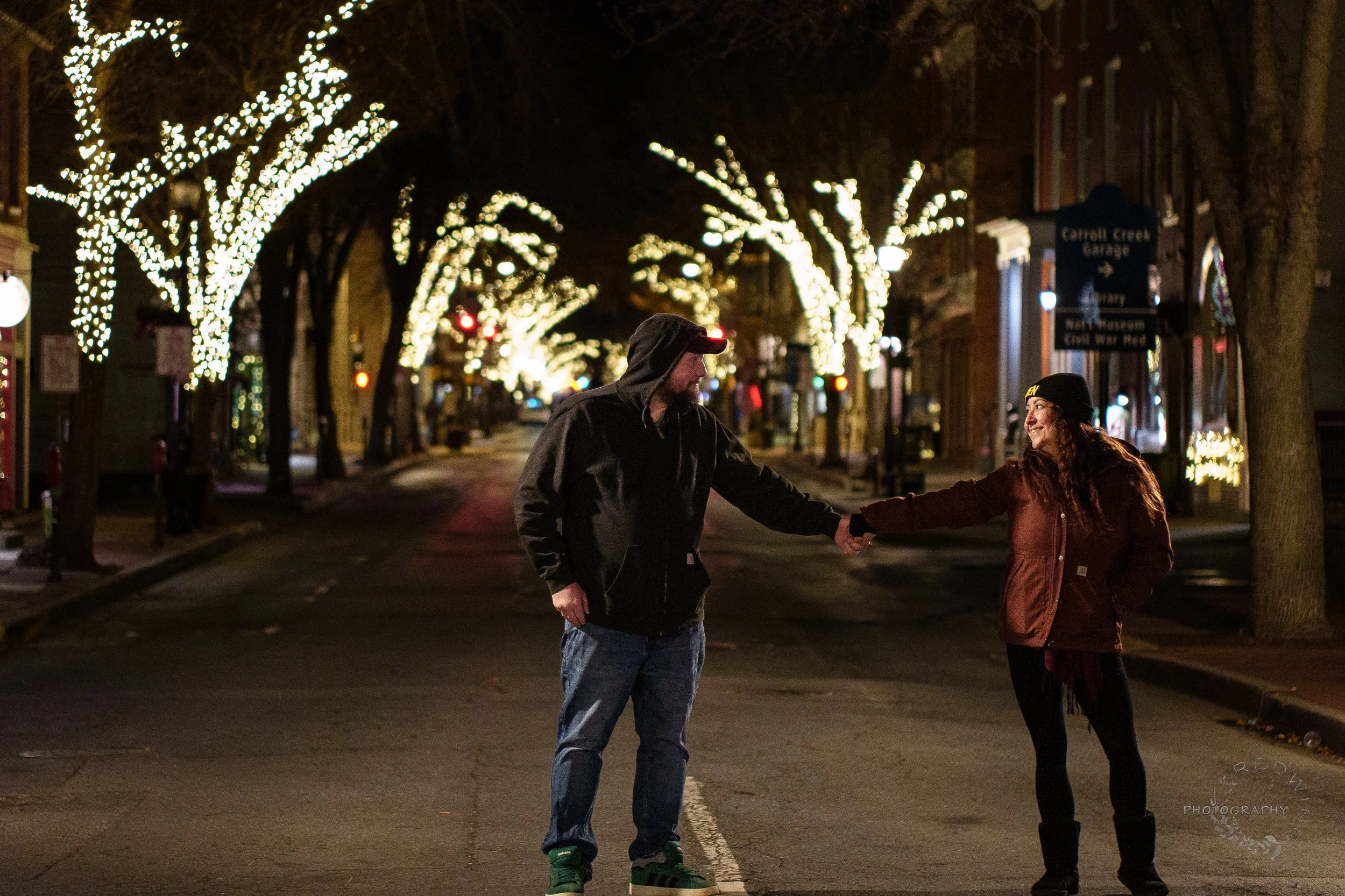 A man and woman shaking hands in the middle of a lit street at night, decorated with Christmas lights on trees.