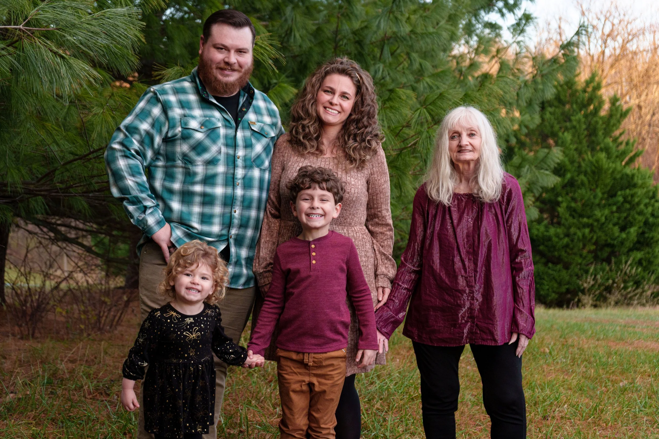 A multigenerational family standing outdoors in front of trees and foliage, smiling for a photo. The group includes two adult men, two adult women, and two children, all wearing casual clothing.
