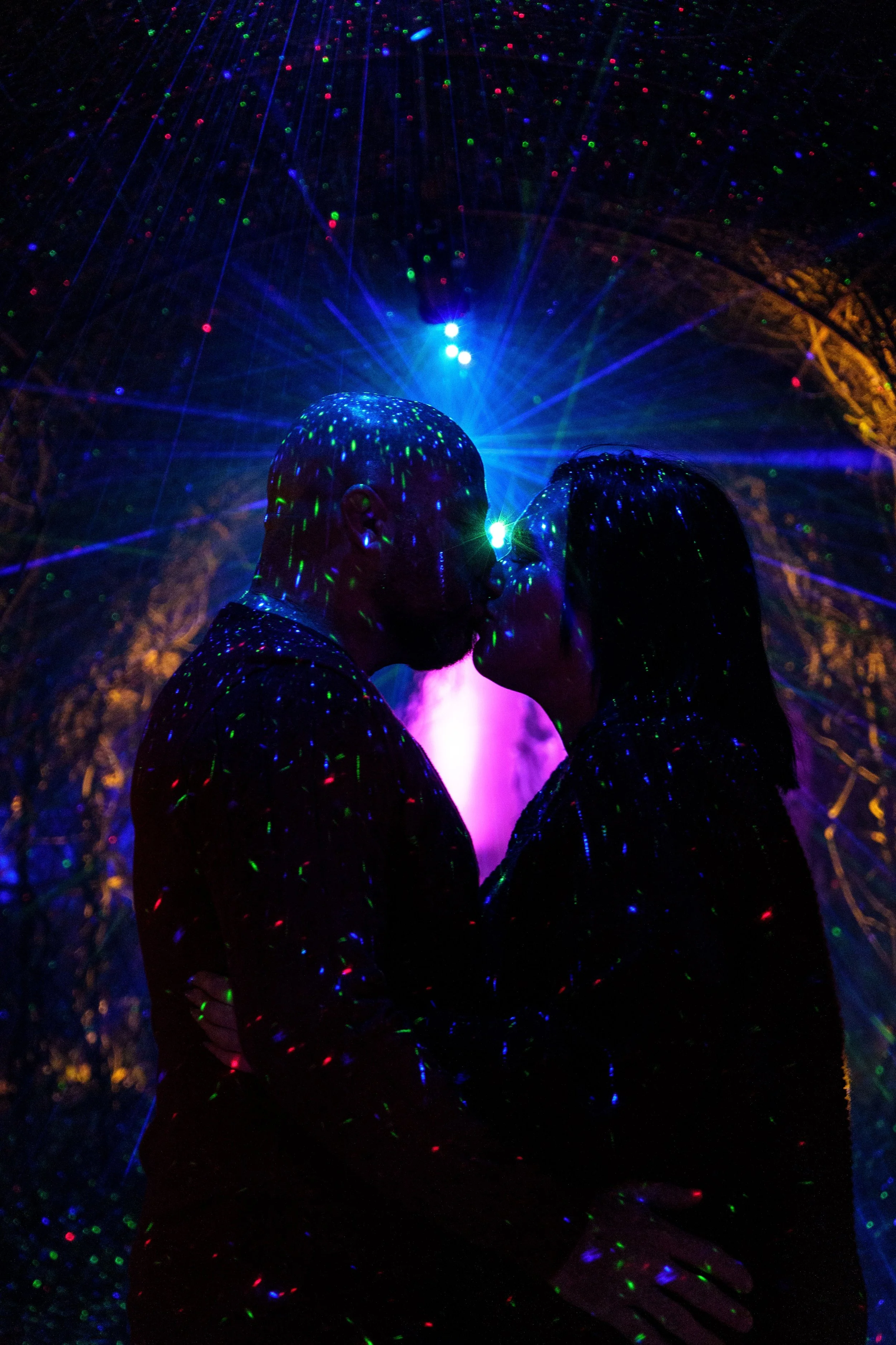 Silhouettes of a man and woman sharing a kiss in a dark environment illuminated by colorful laser lights.